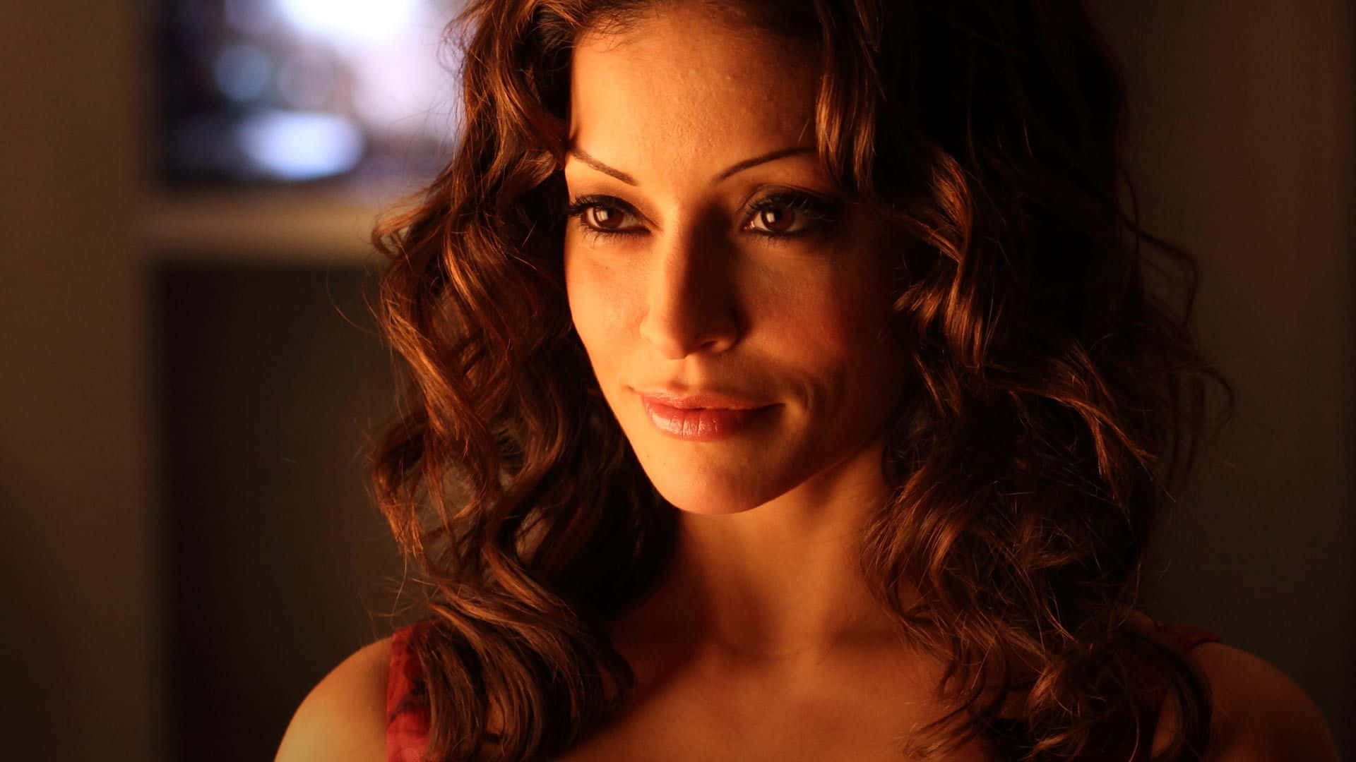 Emmanuelle Vaugier 桌布 壁紙 — 一位擁有棕色捲髮的女性在昏暗的房間裡，在溫暖的金色燈光下溫柔地凝視著鏡頭。
