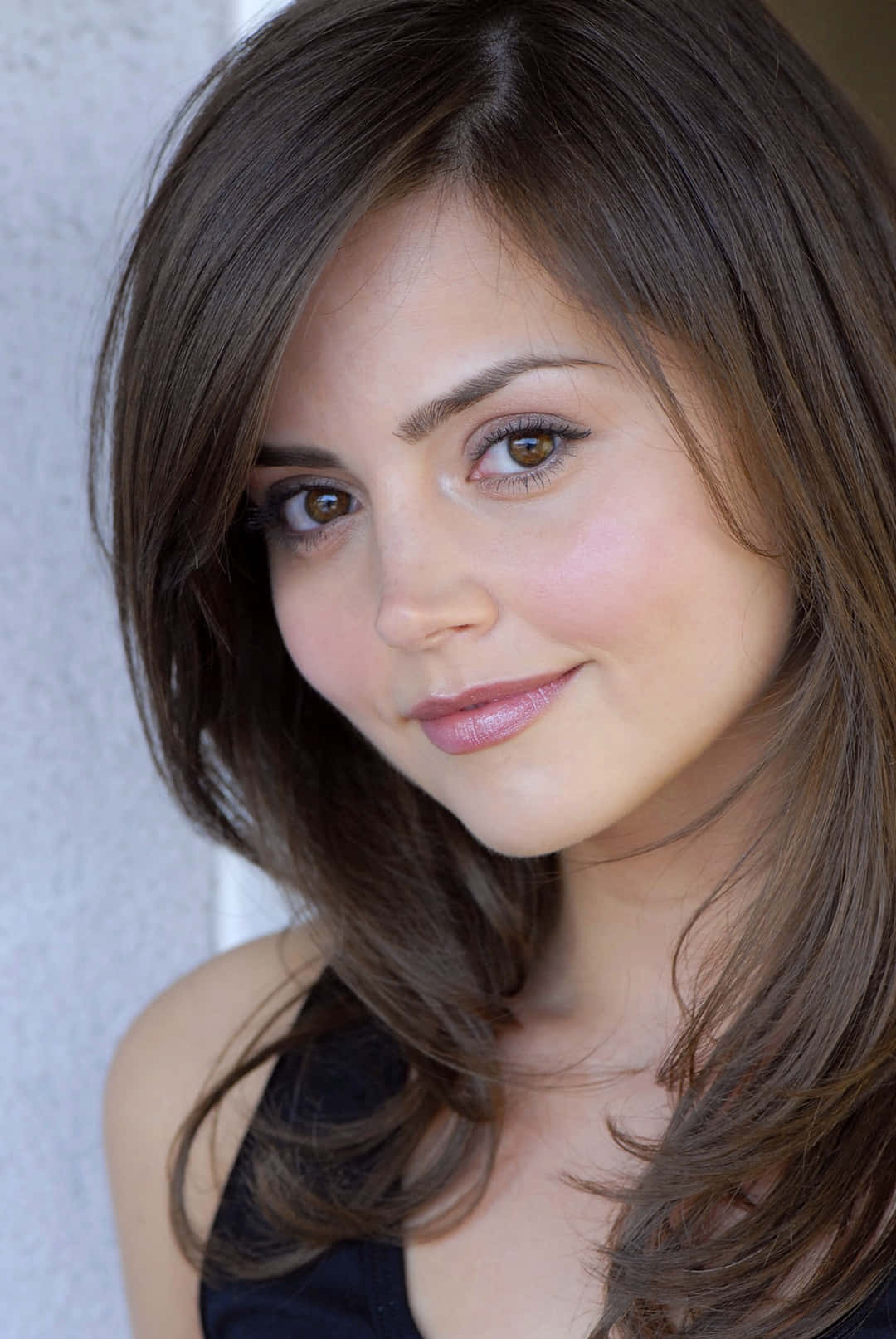 Jenna Coleman 桌布 壁紙 — 一位擁有棕色頭髮和溫暖眼睛的女性，在紋理牆壁前微笑的特寫肖像。