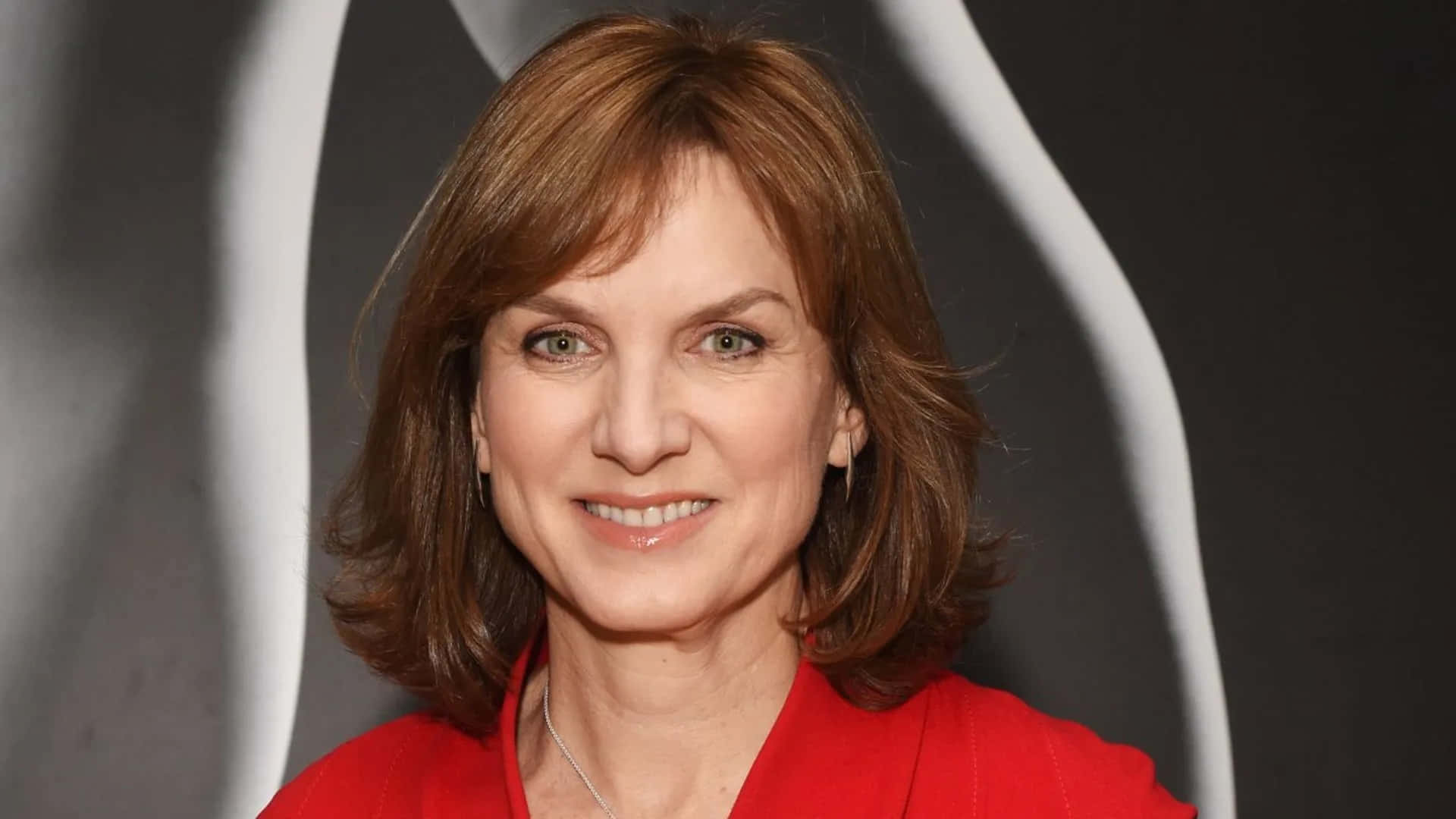 Fiona Bruce 桌布 壁紙 — 一位擁有棕色頭髮和綠色眼睛的女性，穿著紅色上衣，在黑白抽象背景前溫暖地微笑。
