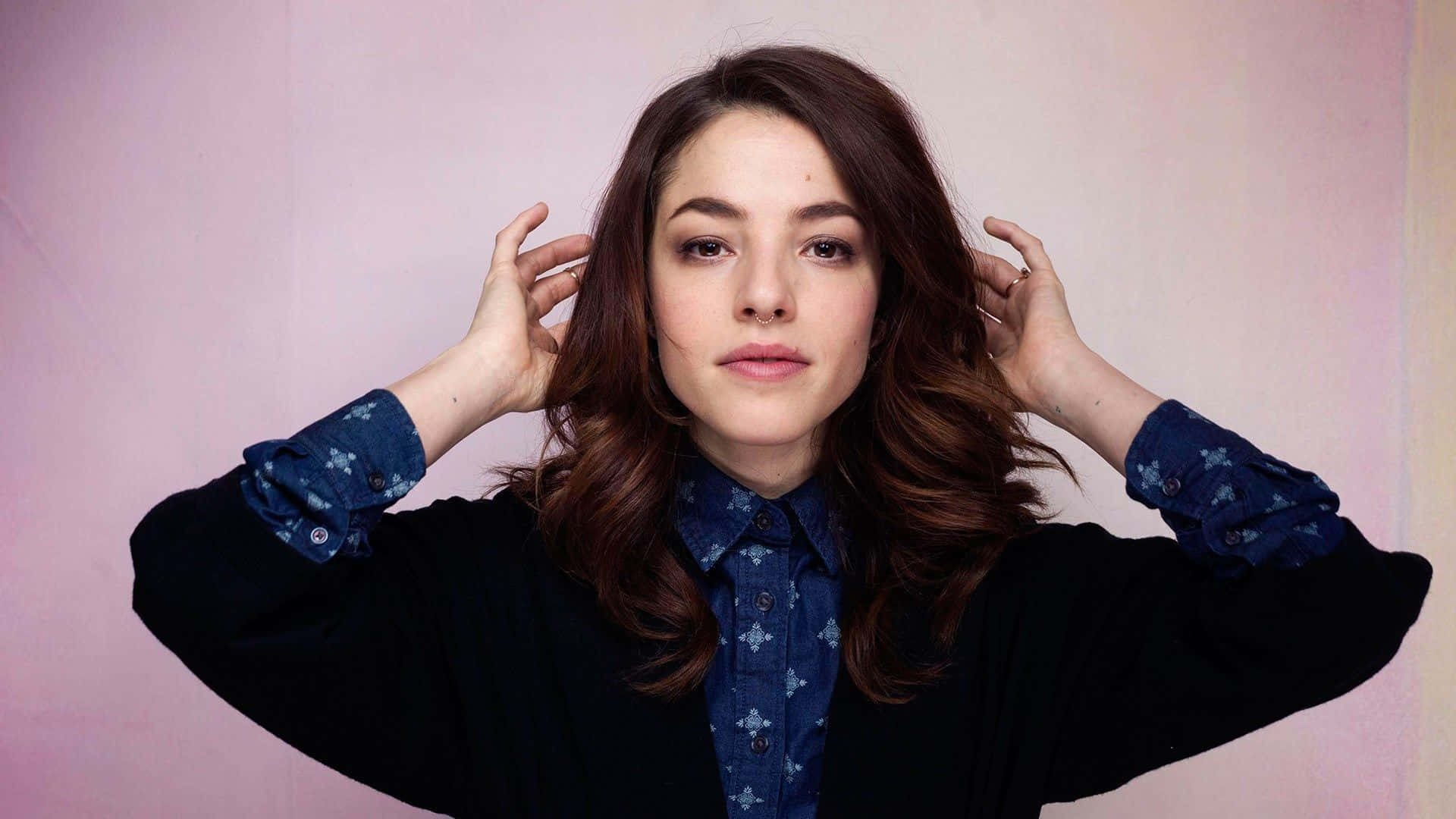 Olivia Thirlby 桌布 壁紙 — 一位擁有波浪棕色長髮的女性，在柔和的粉紅色背景前輕輕觸碰頭部，身穿黑色開襟衫內搭藍色圖案襯衫。