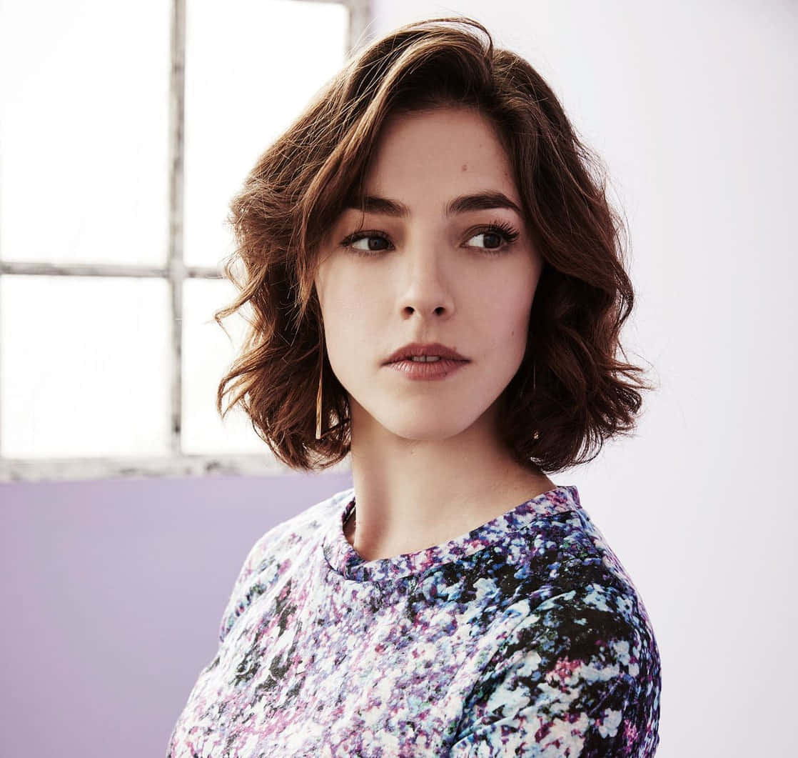 Olivia Thirlby 桌布 壁紙 — 一位擁有波浪棕色長髮的女性，在窗邊穿著花卉圖案上衣，若有所思地望向一旁。