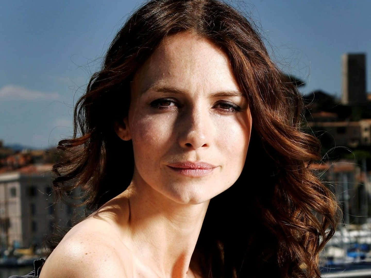 Saffron Burrows 桌布 壁紙 — 一位擁有波浪棕色長髮的女性，在陽光普照的海濱城市背景下平靜地凝視著鏡頭。