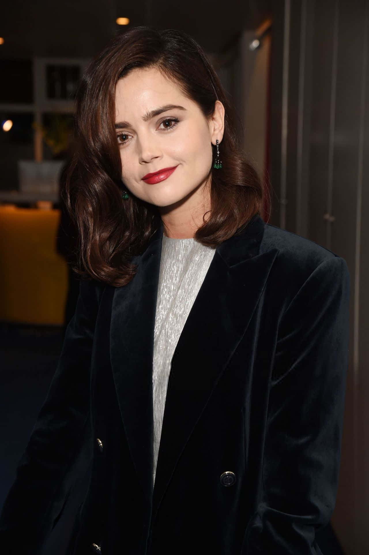Jenna Coleman 桌布 壁紙 — 一位擁有波浪棕色頭髮和紅色唇膏的女性，穿著黑色天鵝絨西裝外套搭配銀色上衣，溫柔地微笑。