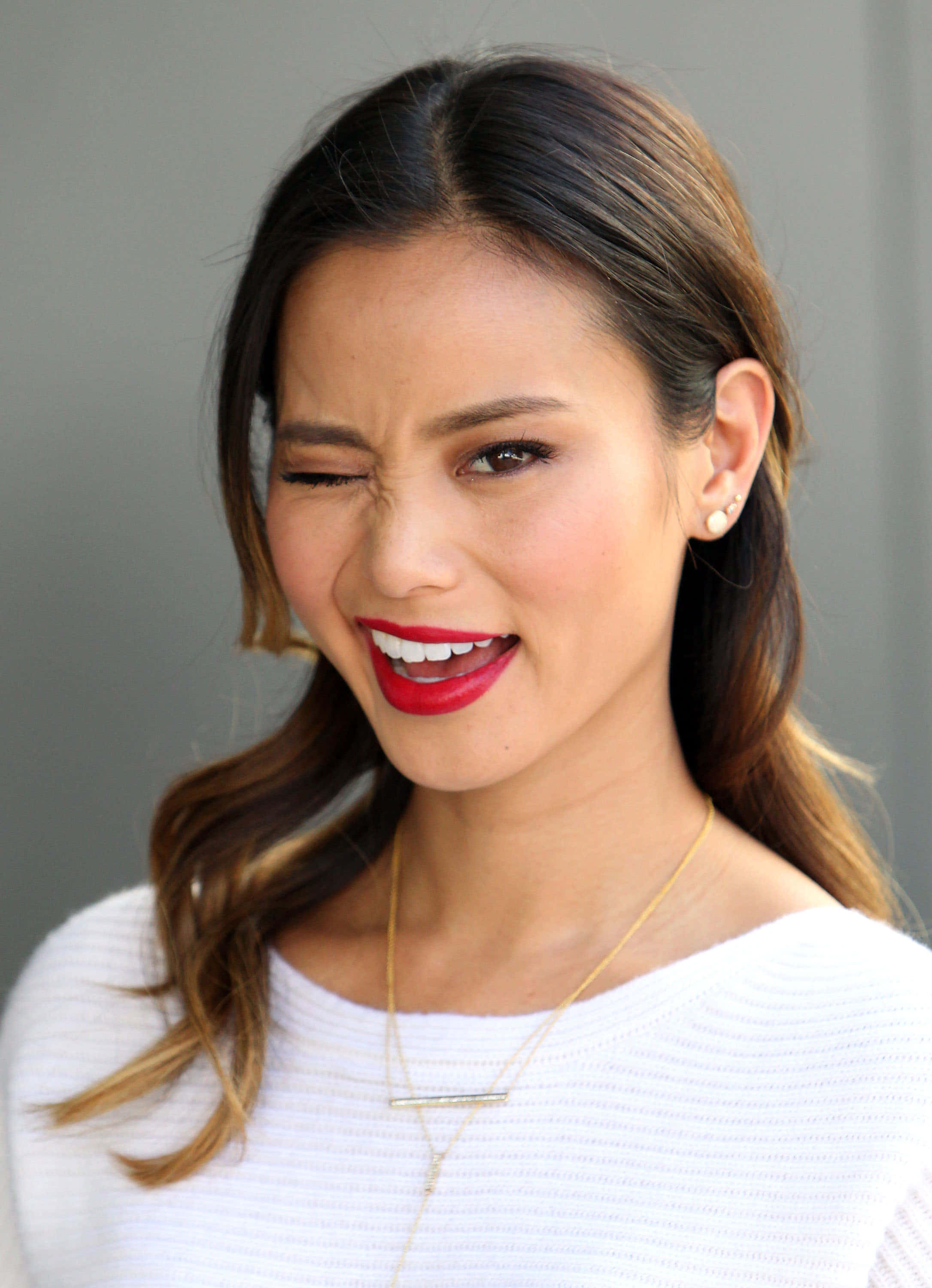 Jamie Chung 桌布 壁紙 — 一位擁有波浪棕色頭髮和鮮豔紅色唇膏的女性，在純灰色背景前閉著眼睛大笑。