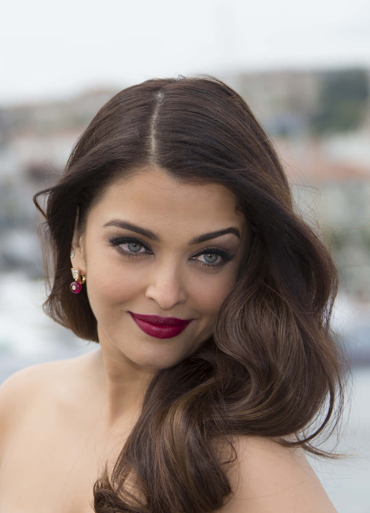 Aishwarya Rai 桌布 壁紙 — 一位擁有波浪棕色頭髮和鮮豔紅色唇膏的女性，在模糊的城市背景下溫柔微笑。