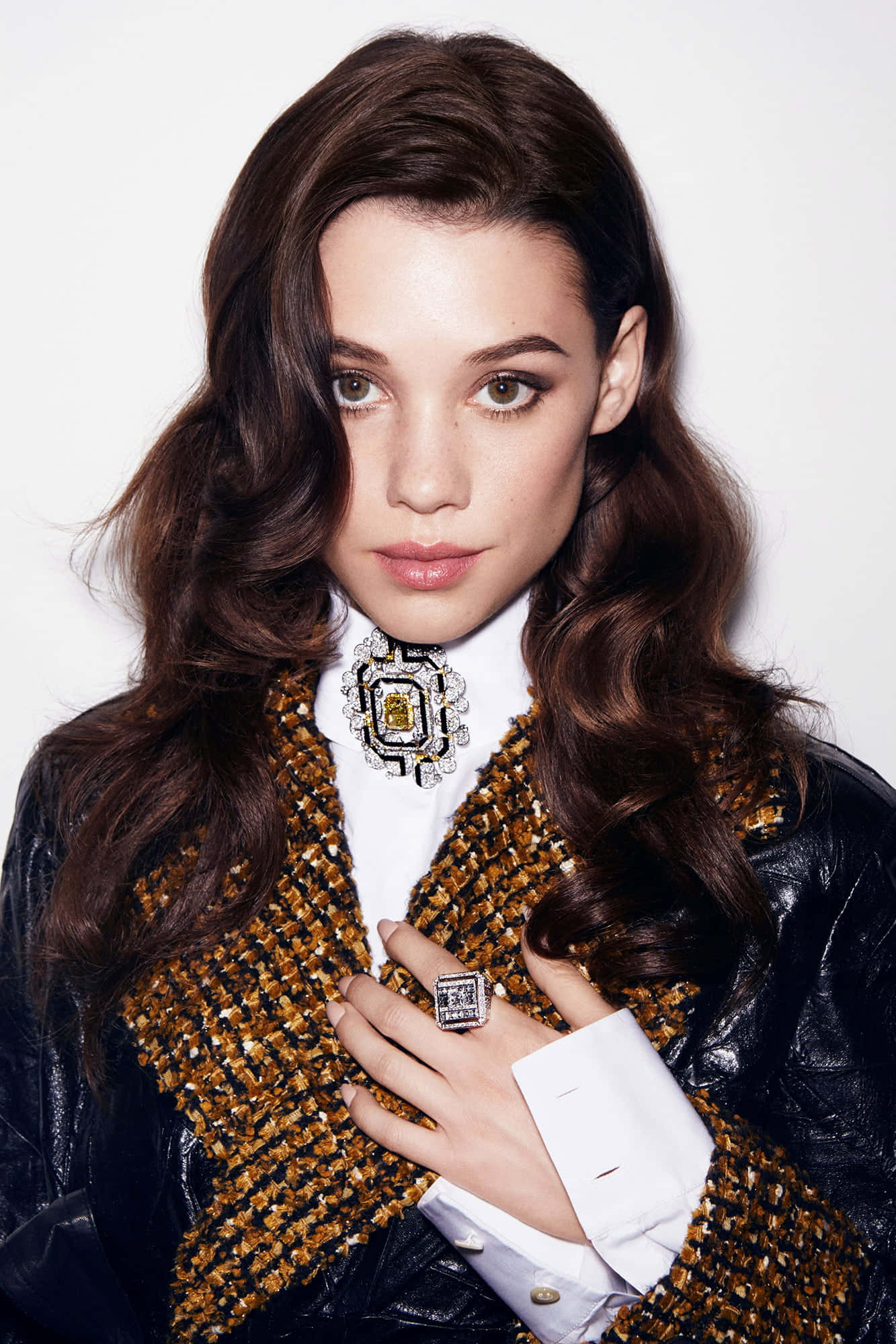 Astrid Berges Frisbey 桌布 壁紙 — 一位擁有波浪棕色頭髮的女性，身穿黑色皮褸，內搭金色粗花呢背心和白色襯衫，佩戴大型幾何胸針和相配的戒指。