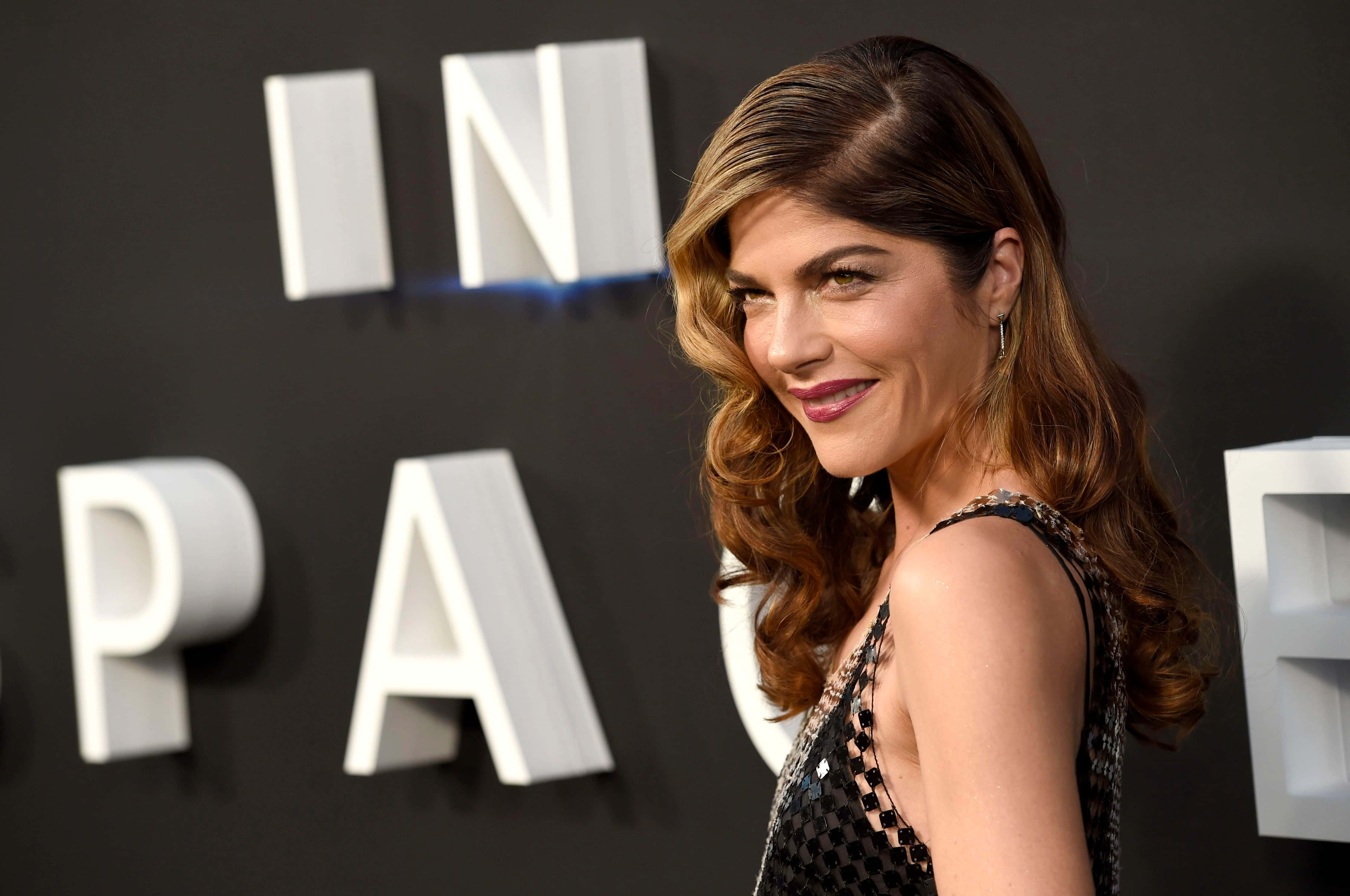 Selma Blair 桌布 壁紙 — 一位擁有波浪棕色頭髮的女性在紅地毯活動中對著鏡頭微笑，身後有大型白色字母拼出「IN SPACE」。