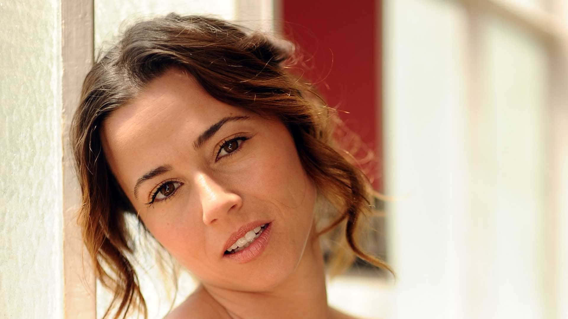 linda cardellini 桌布 壁紙 — 一位擁有波浪棕色頭髮的女性靠在白色牆壁上，身後有紅色門框，溫柔地凝視著鏡頭。