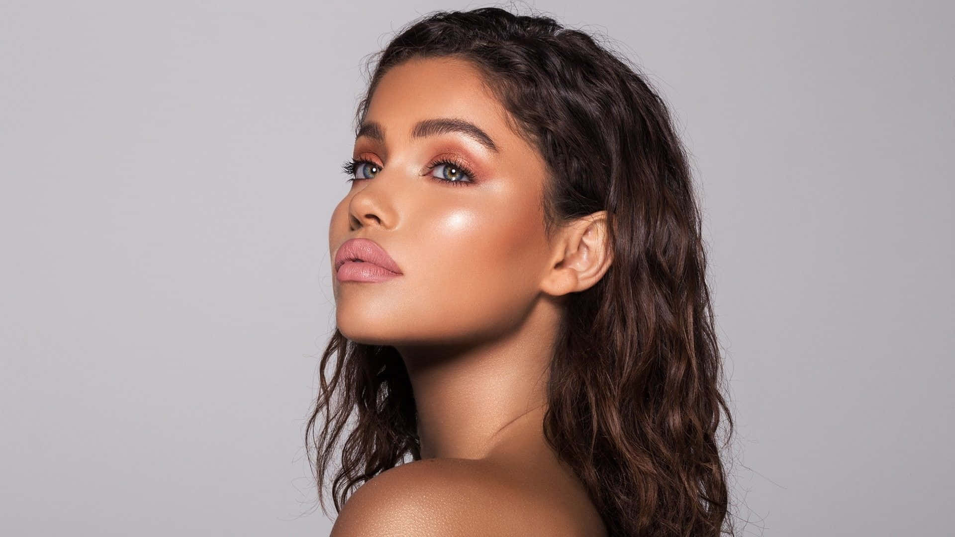 Kylie Jenner 桌布 壁紙 — 一位擁有波浪深色頭髮和發光妝容的女性，在柔和的灰色工作室背景下向上凝視。