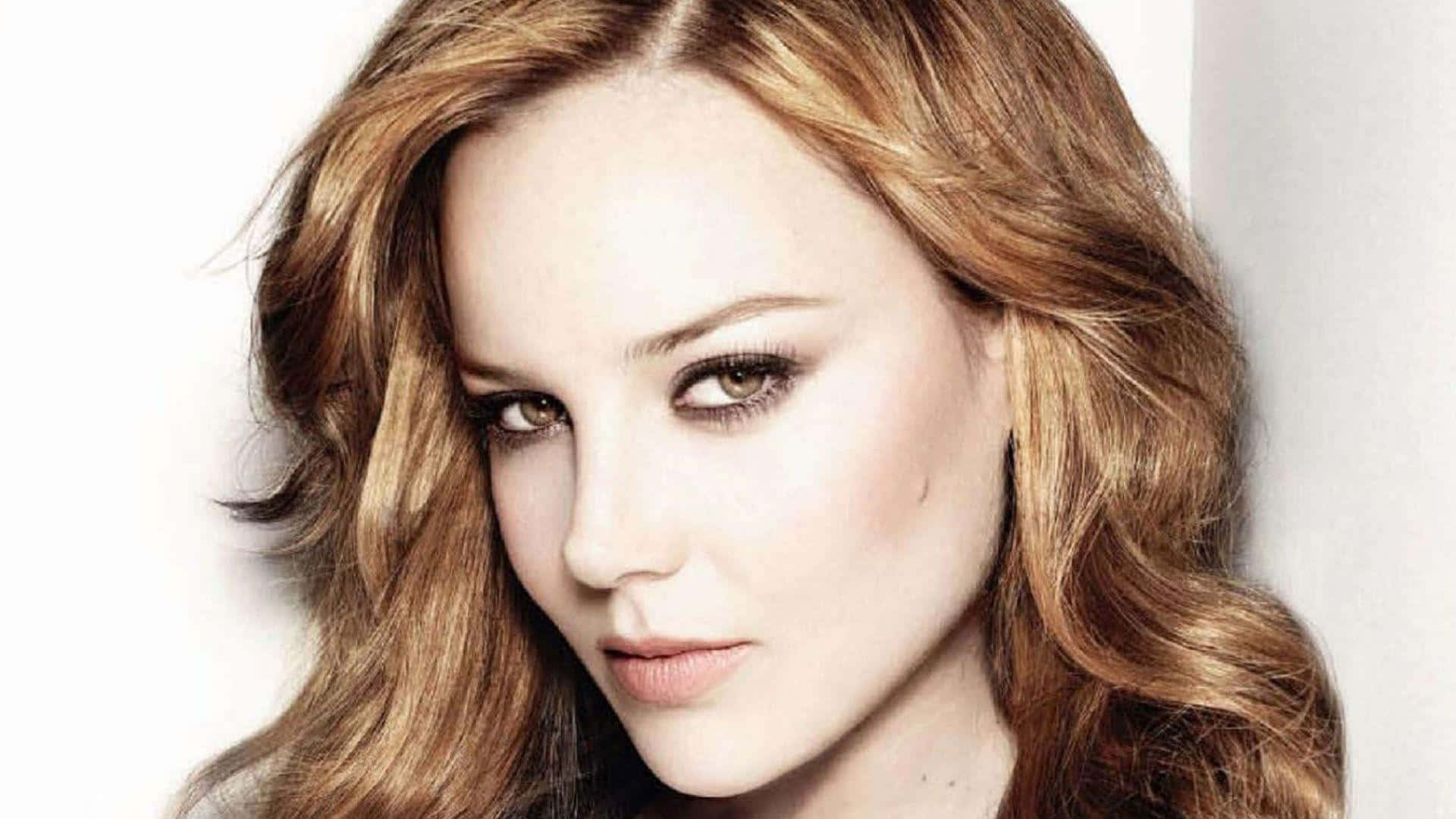 Abbie Cornish 桌布 壁紙 — 一位擁有波浪金色棕色頭髮和煙燻妝眼睛的女性，在純白色背景下的特寫肖像。