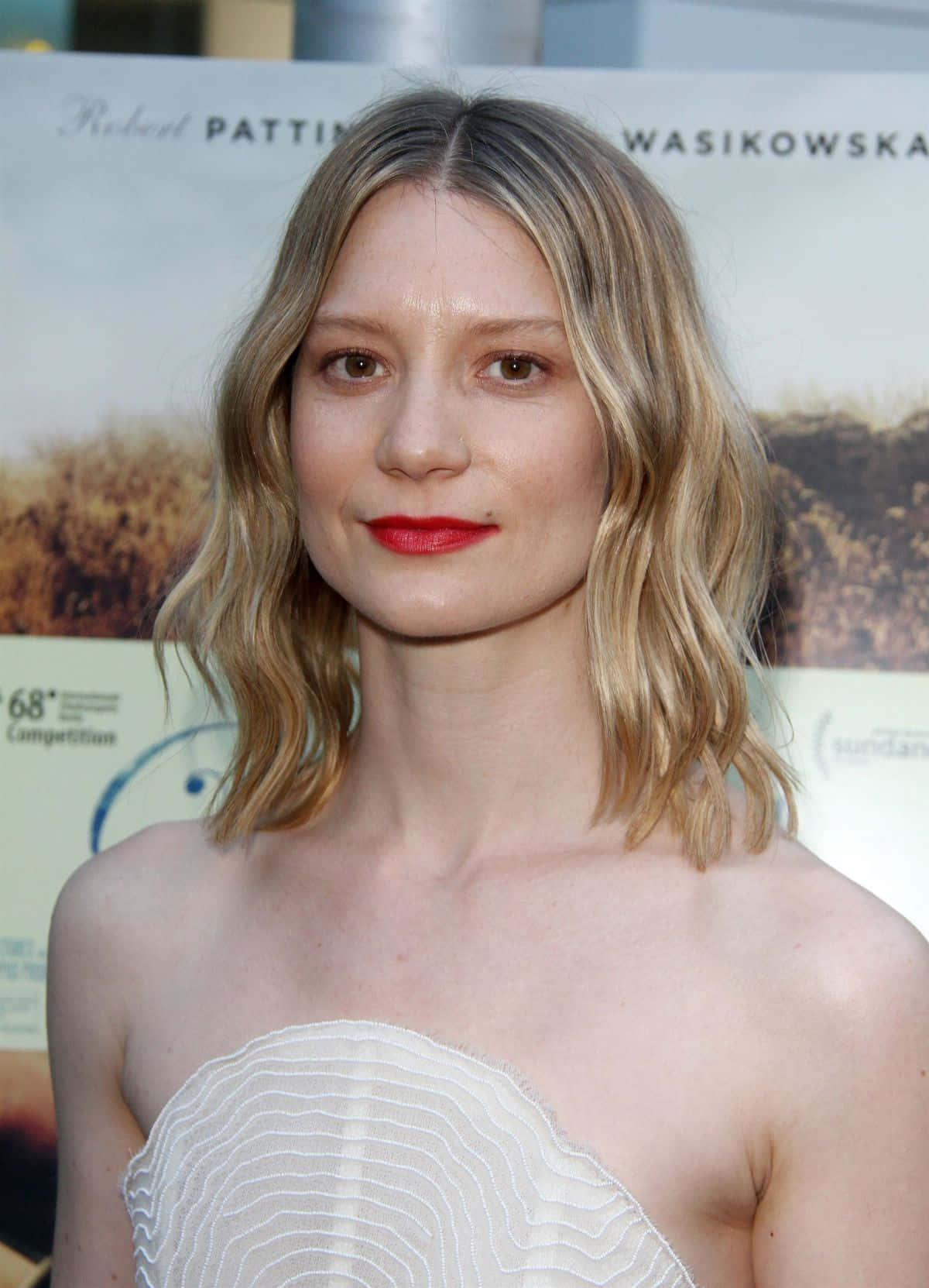 Mia Wasikowska 桌布 壁紙 — 一位擁有波浪金髮和紅唇的女性在電影節活動中穿著白色質感露肩連衣裙。