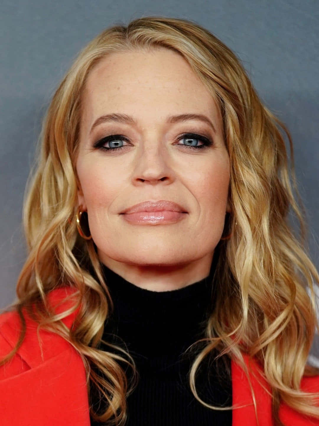 Jeri Ryan 桌布 壁紙 — 一位擁有波浪金髮和藍眼睛的女性特寫肖像，穿著紅色外套和黑色高領毛衣，背景為灰色。