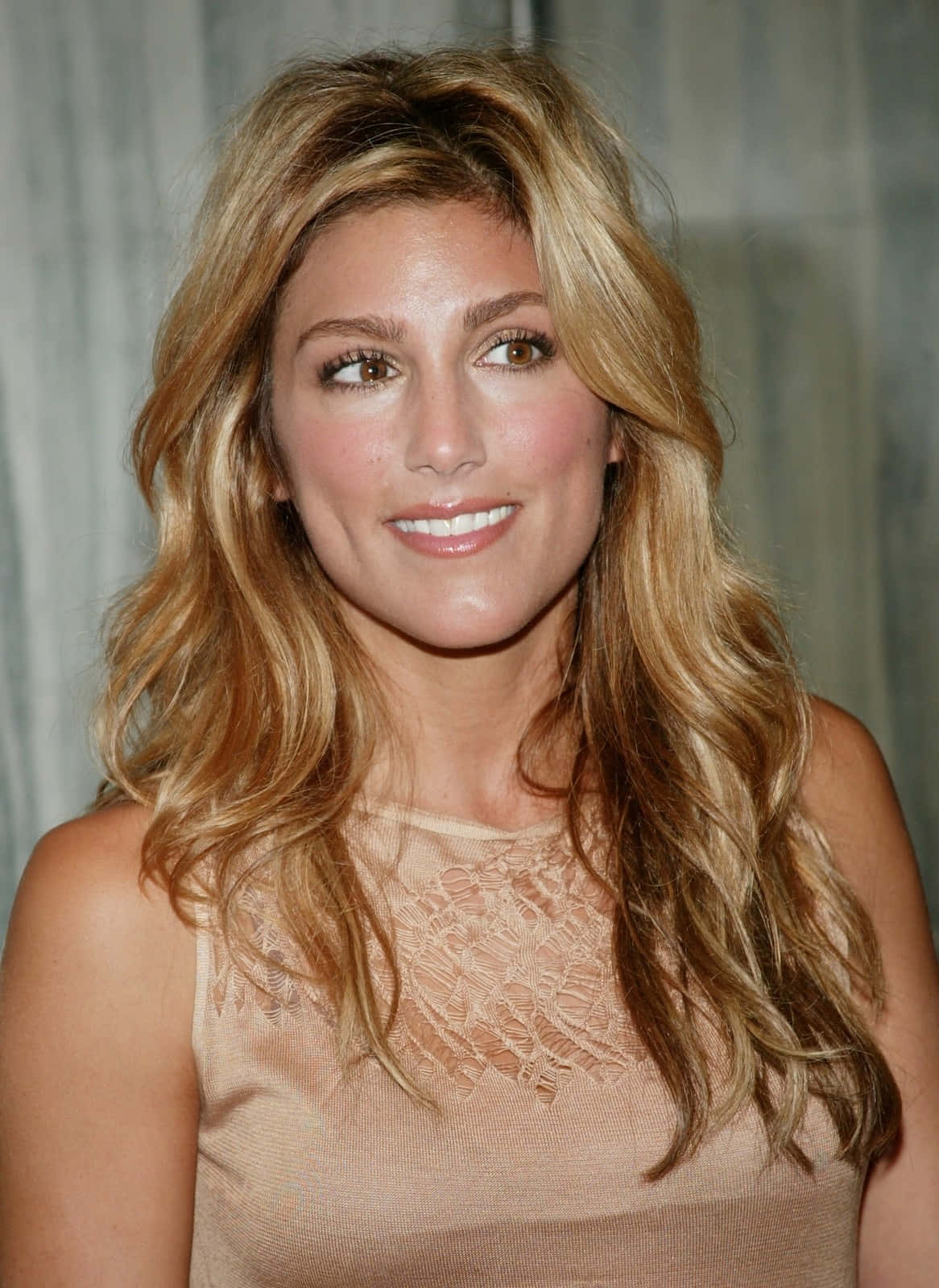 Jennifer Esposito 桌布 壁紙 — 一位擁有波浪金髮的女性，穿著米色蕾絲上衣，在柔和的灰色窗簾背景下溫暖地微笑。