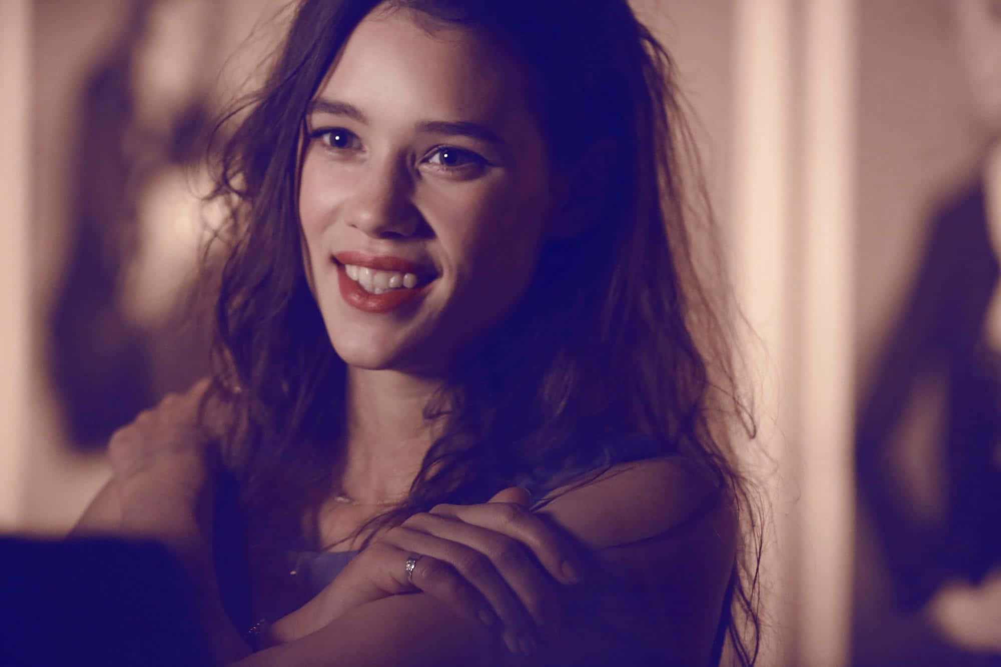 Astrid Berges Frisbey 桌布 壁紙 — 一位擁有波浪長髮的女性在昏暗的房間裡，在柔和的紫色和金色燈光下溫暖地微笑。