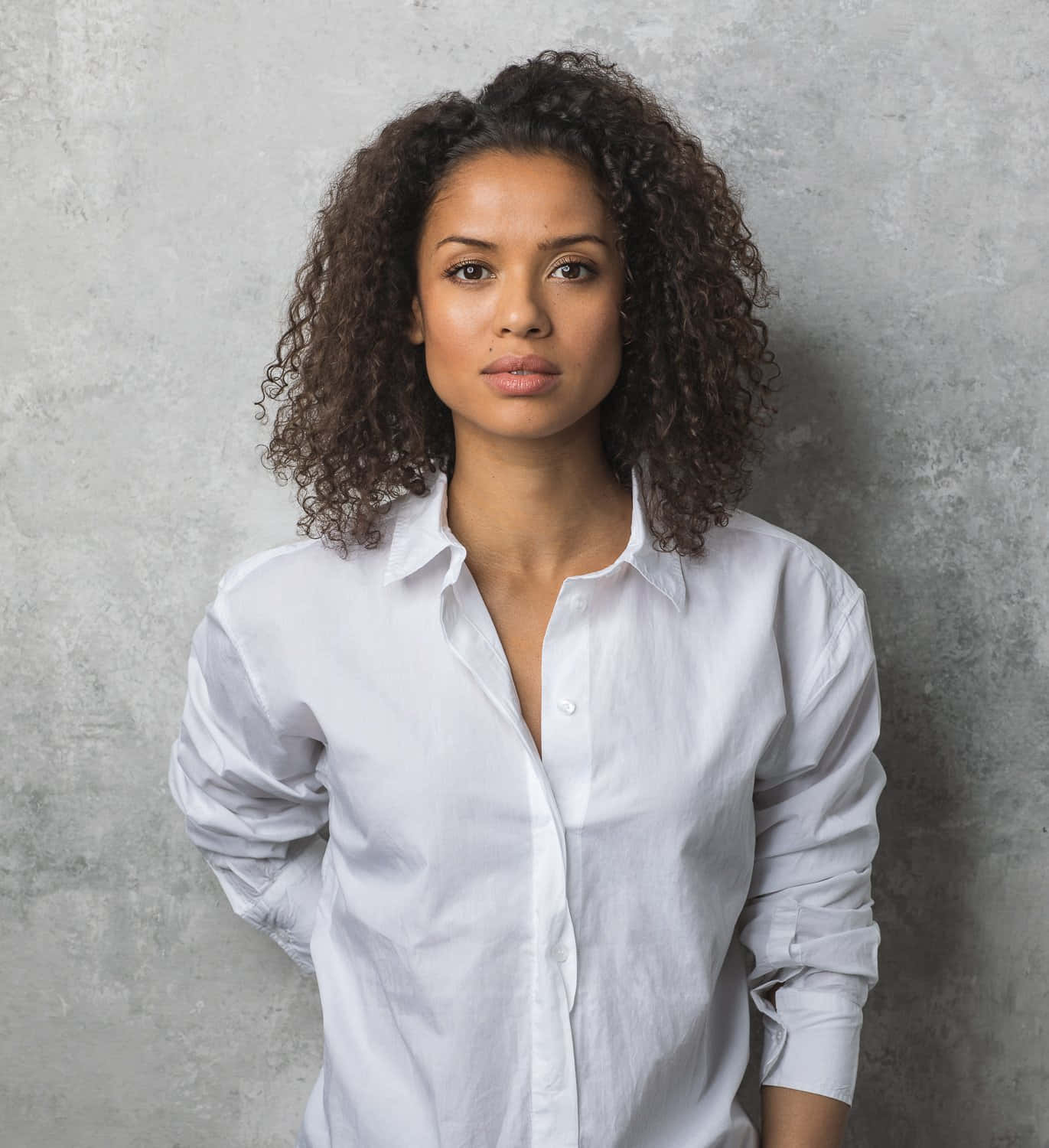 Gugu Mbatha-raw 桌布 壁紙 — 一位擁有深色捲髮的女性，穿著白色鈕扣襯衫，背景是帶有紋理的灰色牆壁。