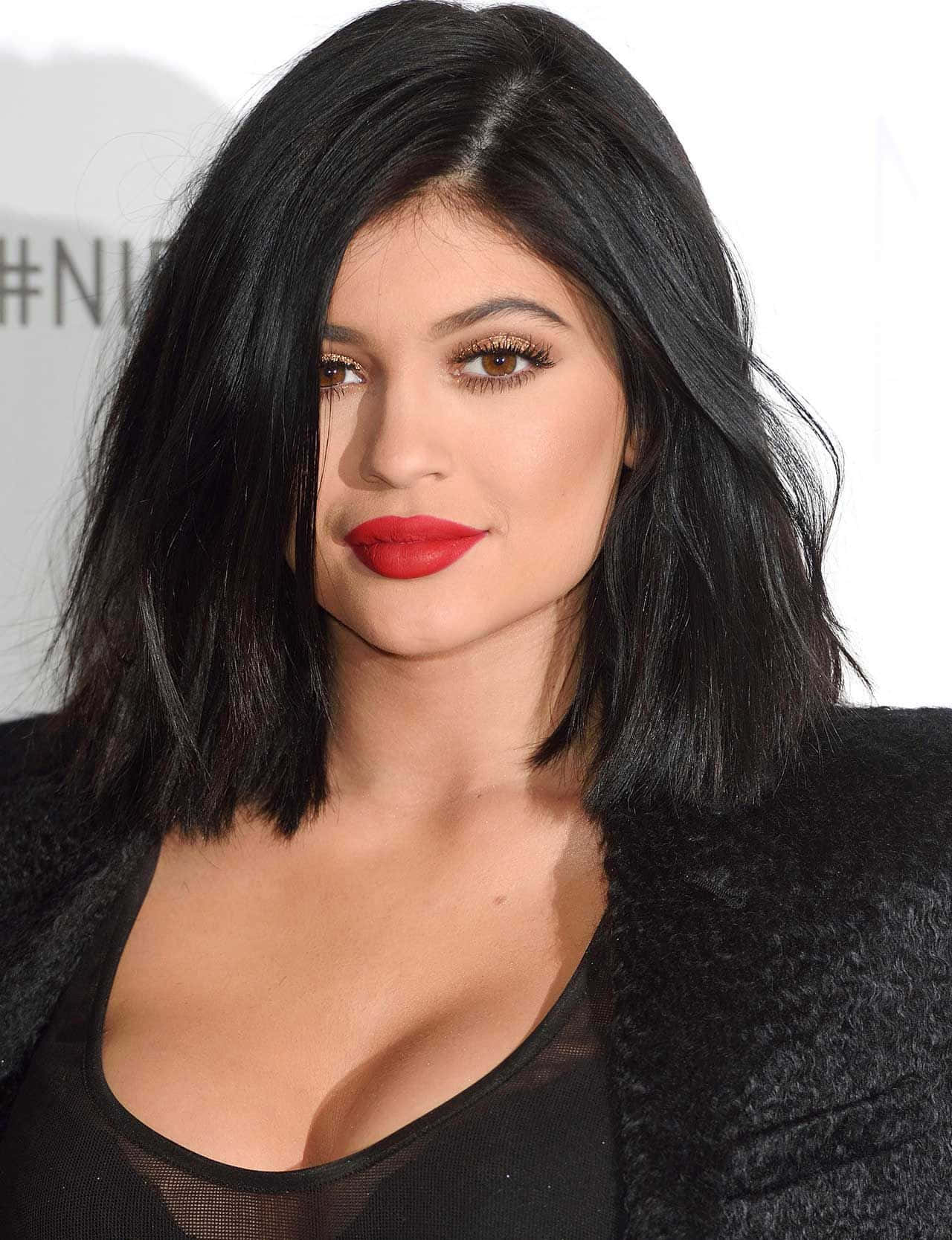 Kylie Jenner 桌布 壁紙 — 一位擁有深色波浪波波頭、大膽紅唇和煙燻妝眼睛的女性穿著黑色紋理外套的特寫肖像。