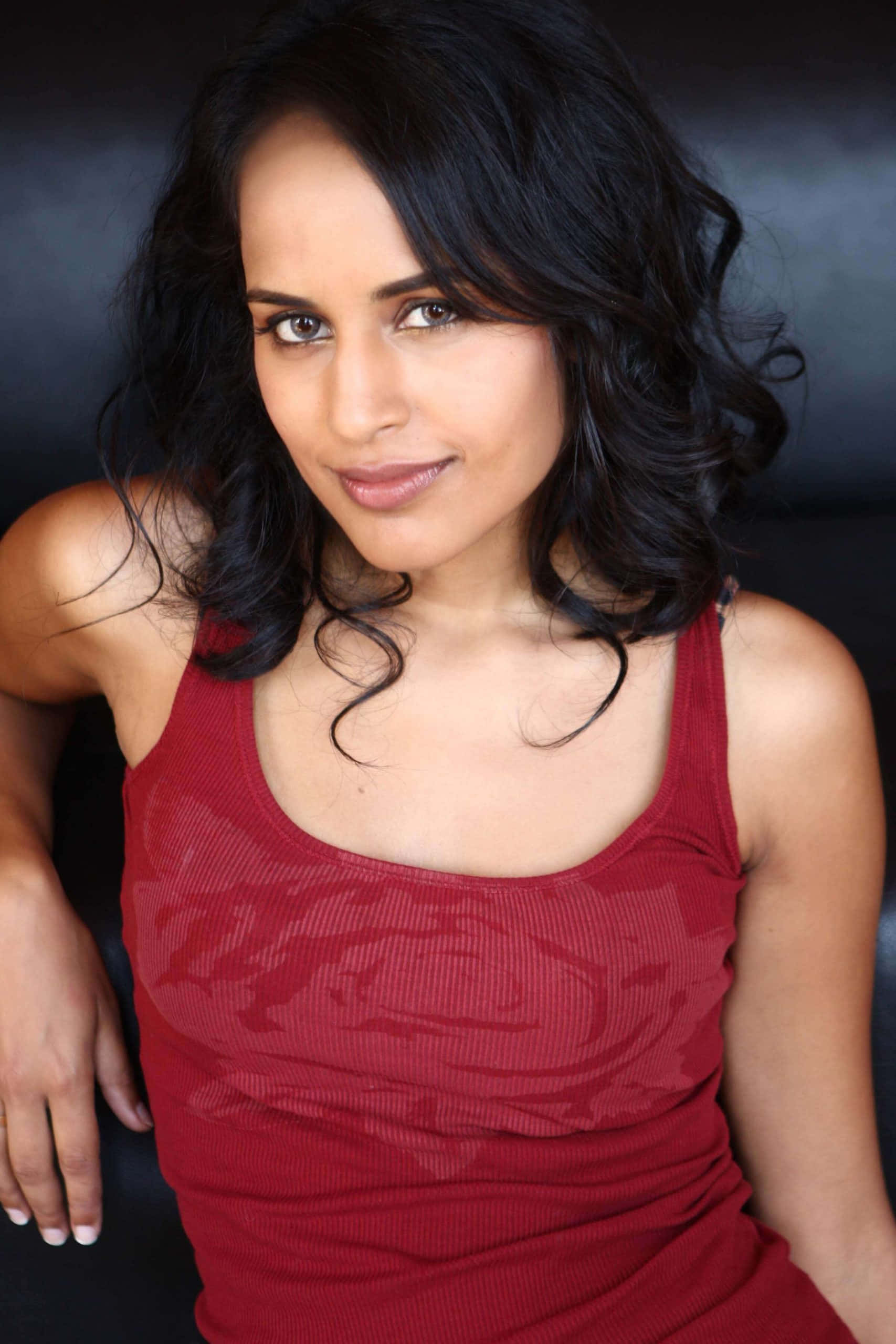 Agam Darshi 桌布 壁紙 — 一位擁有深色波浪長髮、身穿紅色羅紋背心的女性，在深色背景前溫柔微笑。
