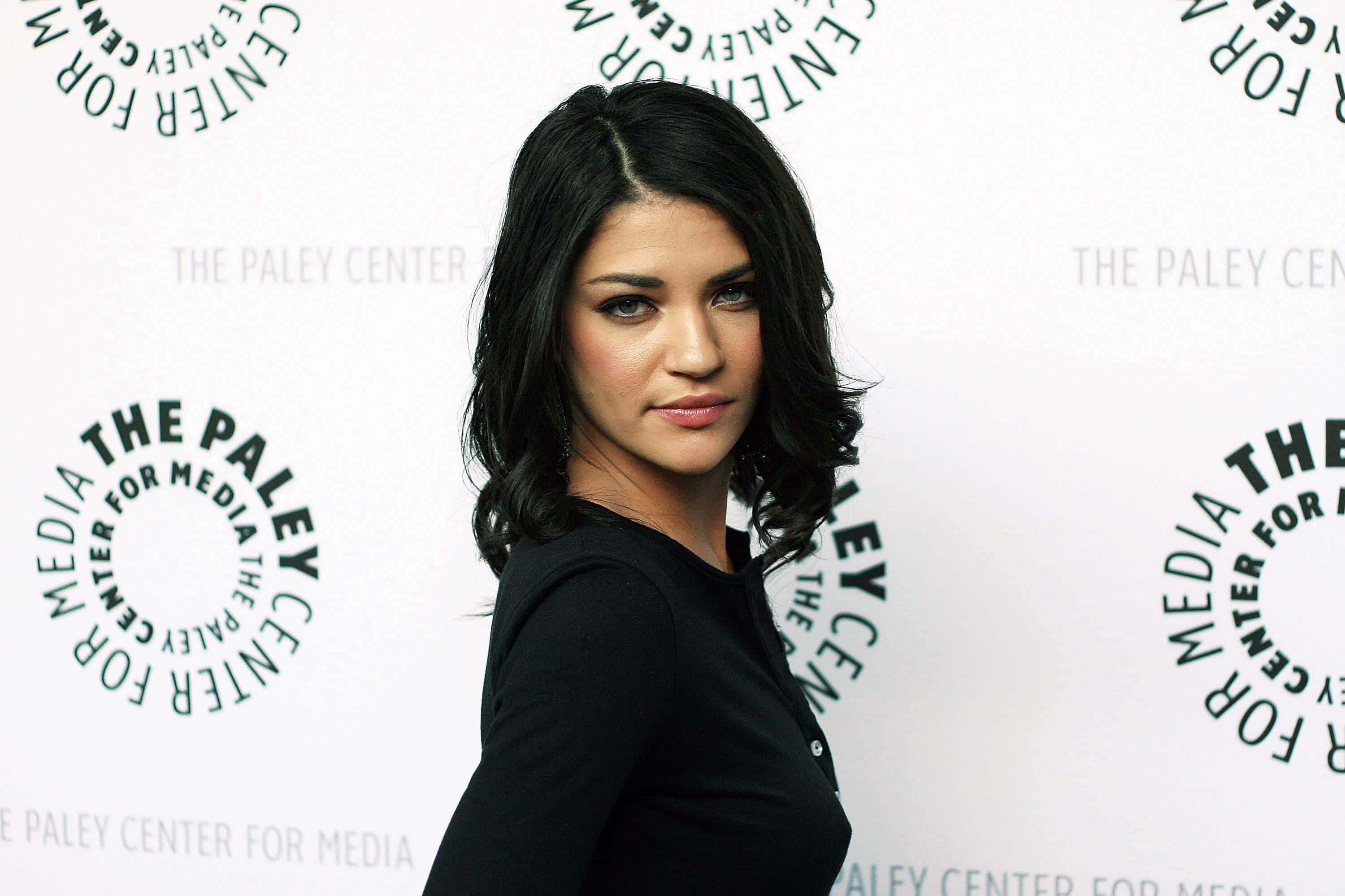 jessica szohr 桌布 壁紙 — 一位擁有深色波浪長髮、身穿黑色上衣的女性，在白色背景前自信地擺姿勢，背景上重複印有綠色「The Paley Center for Media」標誌。