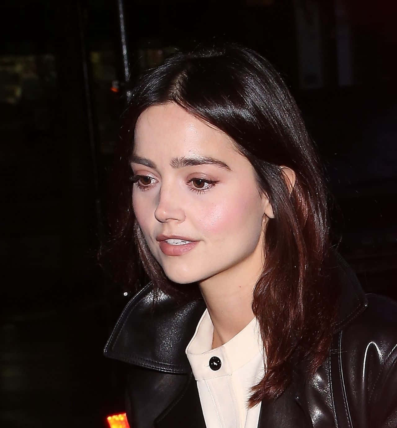 Jenna Coleman 桌布 壁紙 — 一位擁有深色波浪長髮的女性，在深色夜間背景下，身穿黑色皮褸內搭白色襯衫。