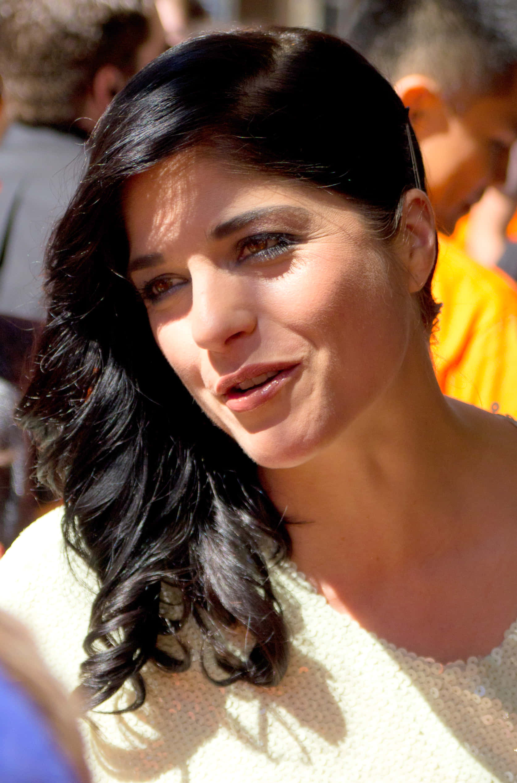Selma Blair 桌布 壁紙 — 一位擁有深色波浪長髮的女性在明亮陽光下穿著淺色上衣溫柔微笑。