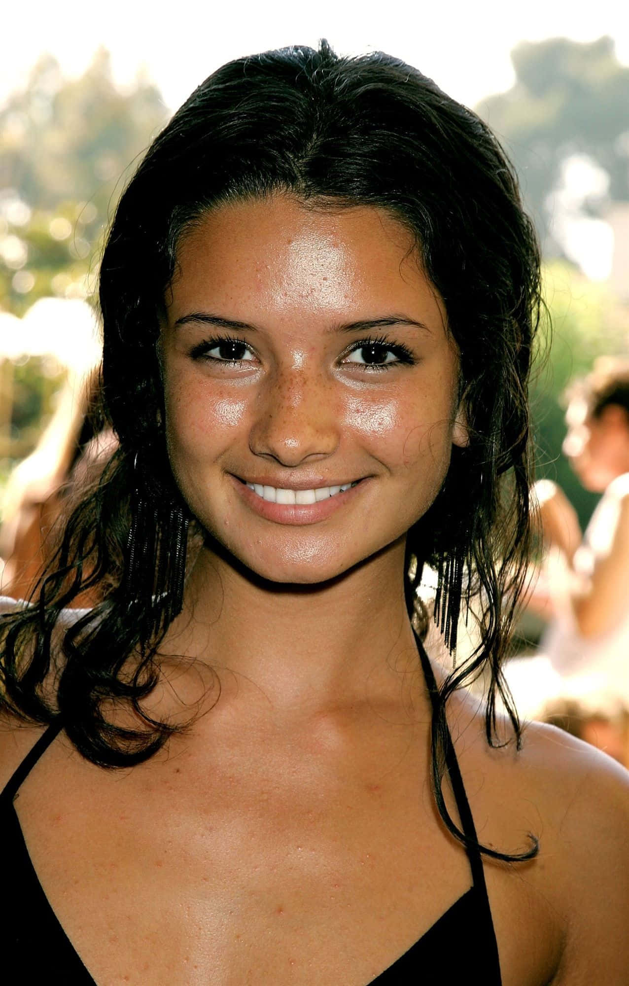 Alice Greczyn 桌布 壁紙 — 一位擁有深色波浪頭髮和雀斑的容光煥發女性，在明亮的戶外背景下溫暖微笑。