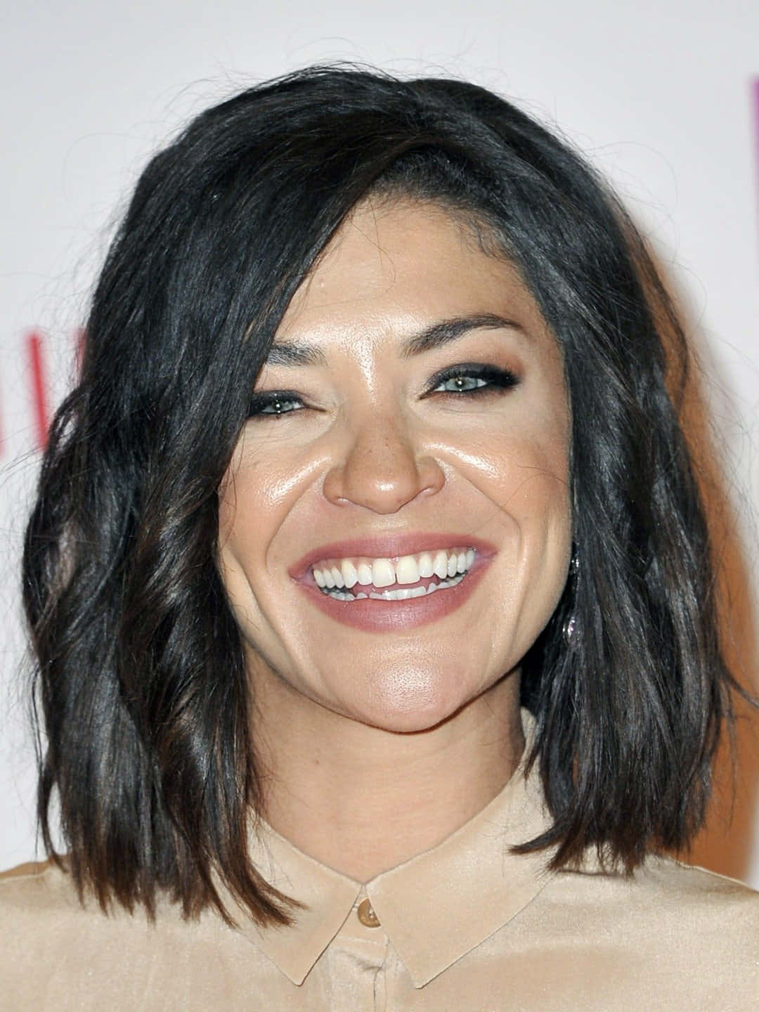 jessica szohr 桌布 壁紙 — 一位擁有深色波浪鮑伯頭和燦爛笑容的女性，穿著米色有領襯衫，背景為白色。