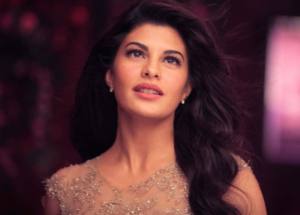 Jacqueline Fernandez 桌布 壁紙 — 一位擁有深色飄逸長髮的迷人女性，身穿閃閃發光的金色連衣裙，在模糊的紫色背景下向上凝視。