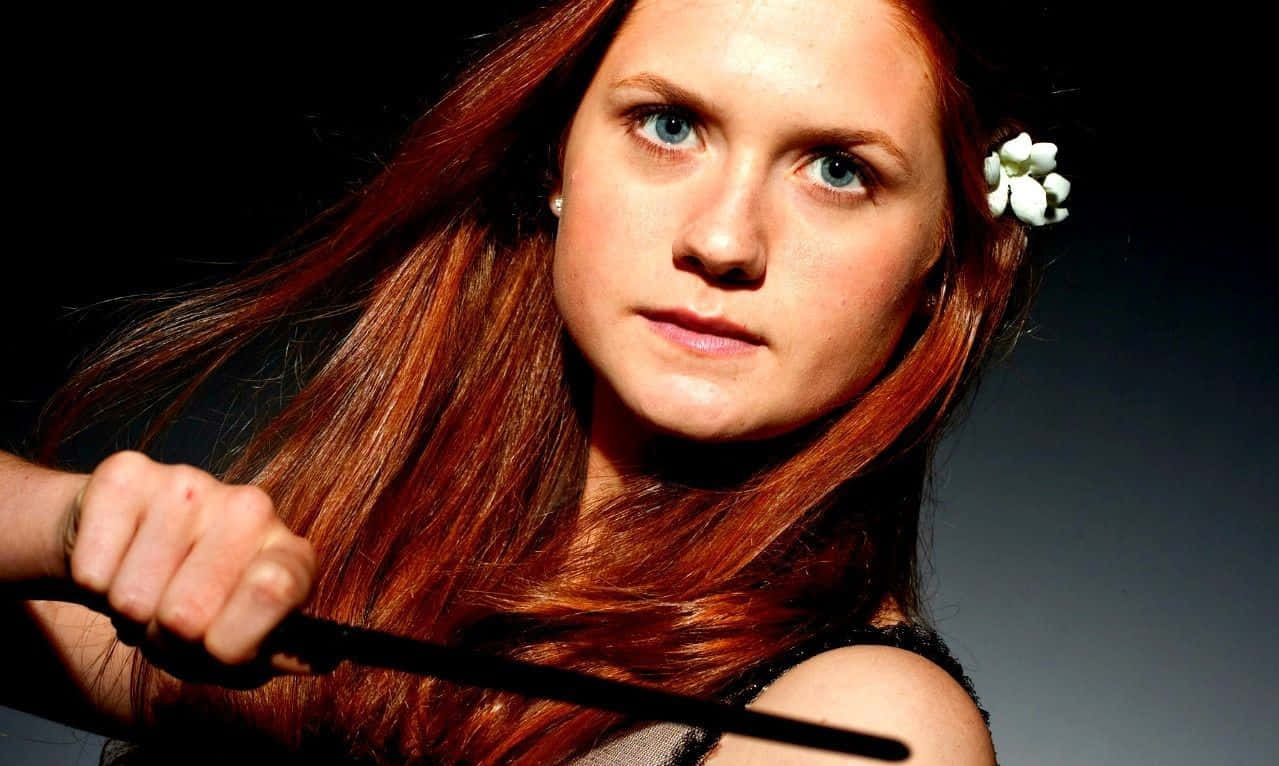 Ginny Weasley 桌布 壁紙 — 一位擁有火紅頭髮和藍色眼睛的女性手持魔杖，一朵白色花朵塞在耳後，背景為深色漸變。