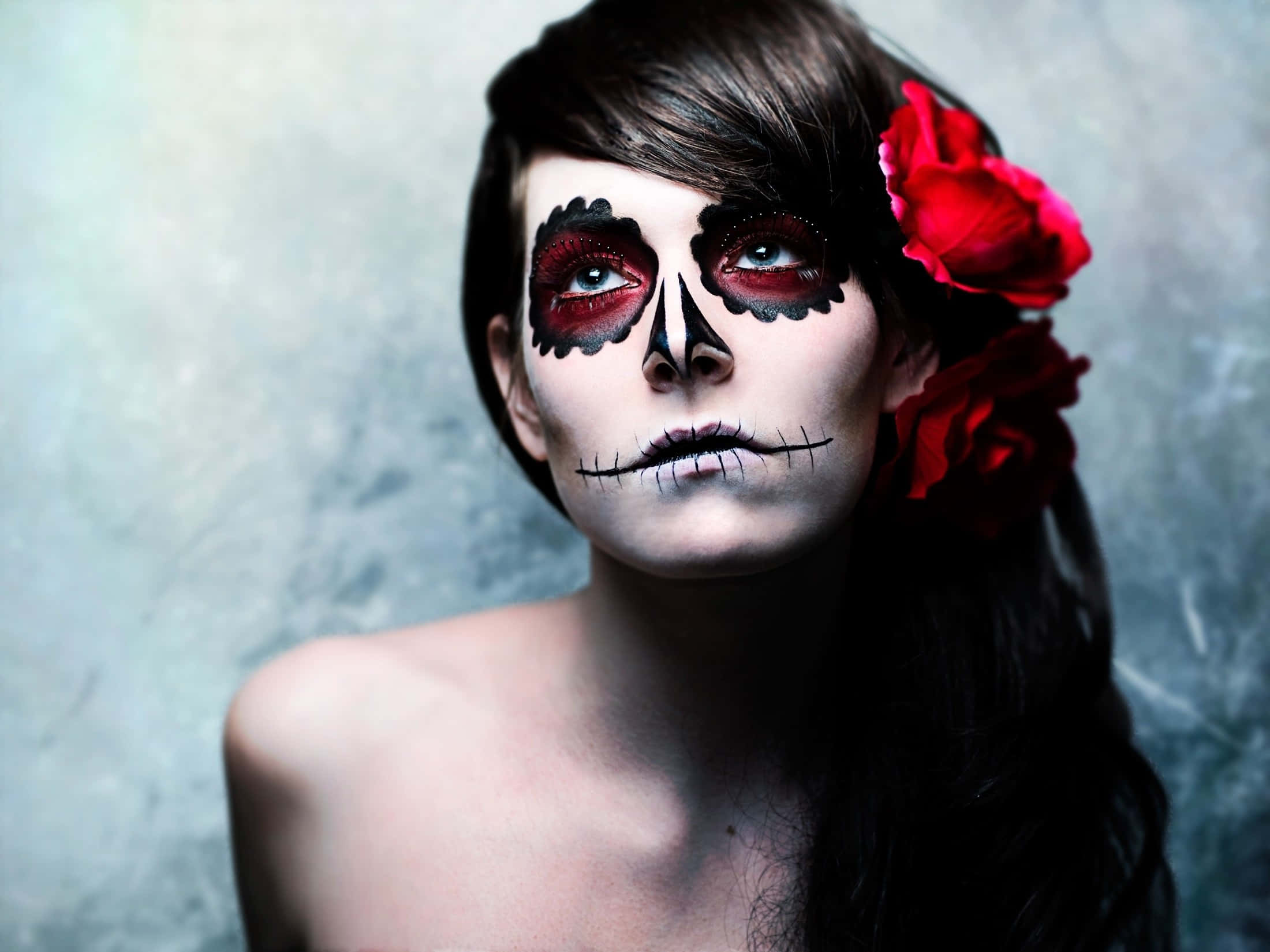 Halloween Makeup 桌布 壁紙 — 一位擁有精緻糖骷髏妝容、頭髮上插著紅玫瑰的女性，直視著帶有紋理的灰色背景。