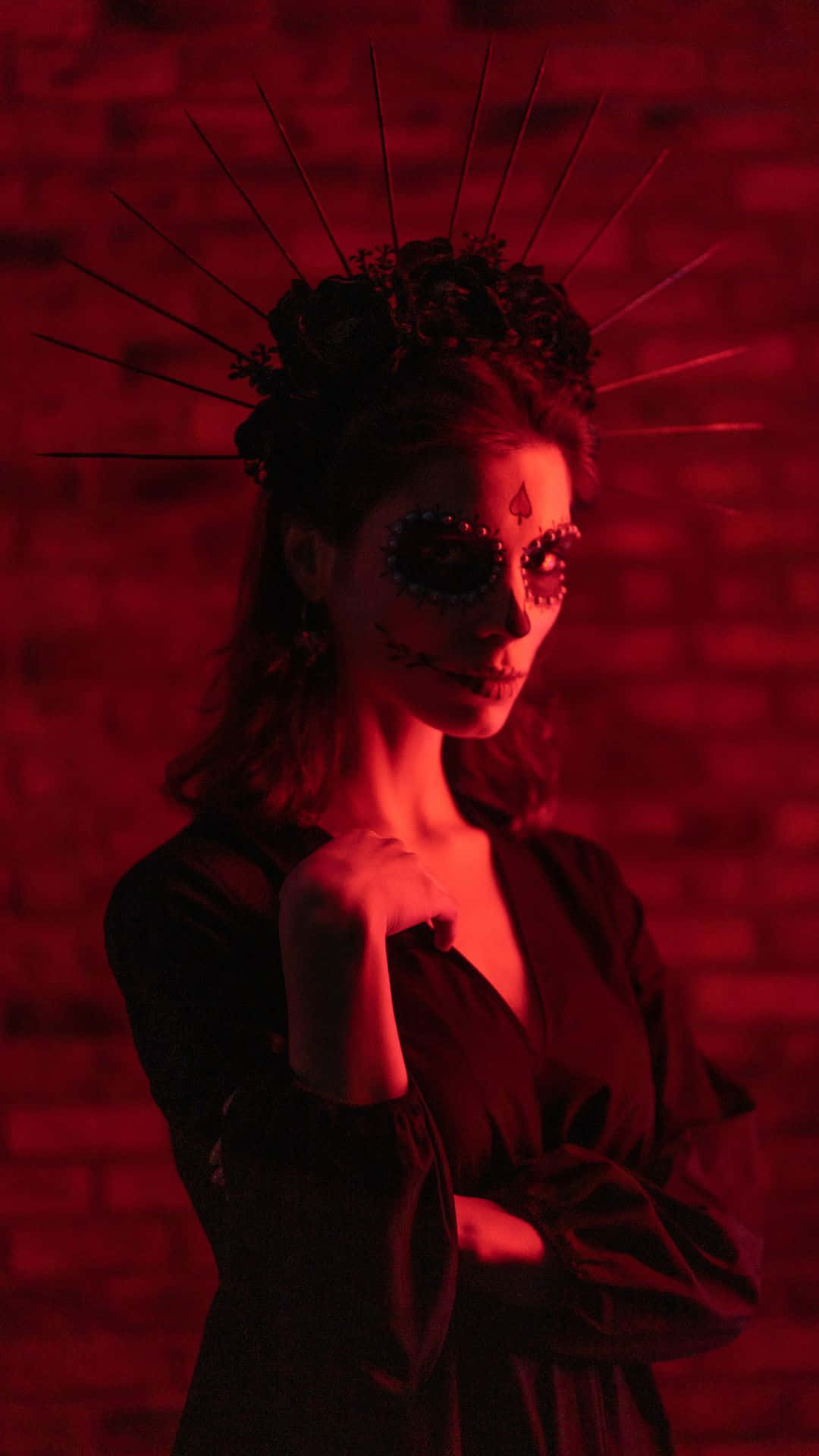 Horror Costumes 桌布 壁紙 — 一位擁有精緻糖骷髏妝容和尖刺皇冠的女性，在戲劇性的紅光下站在紅磚牆前。