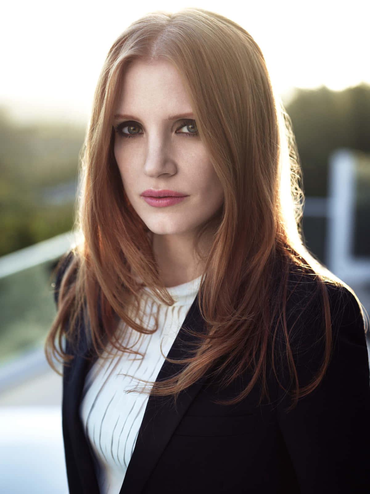 jessica chastain 桌布 壁紙 — 一位擁有紅色長髮、身穿黑色西裝外套的女性站在戶外，金色的陽光突顯了她的頭髮和臉龐。