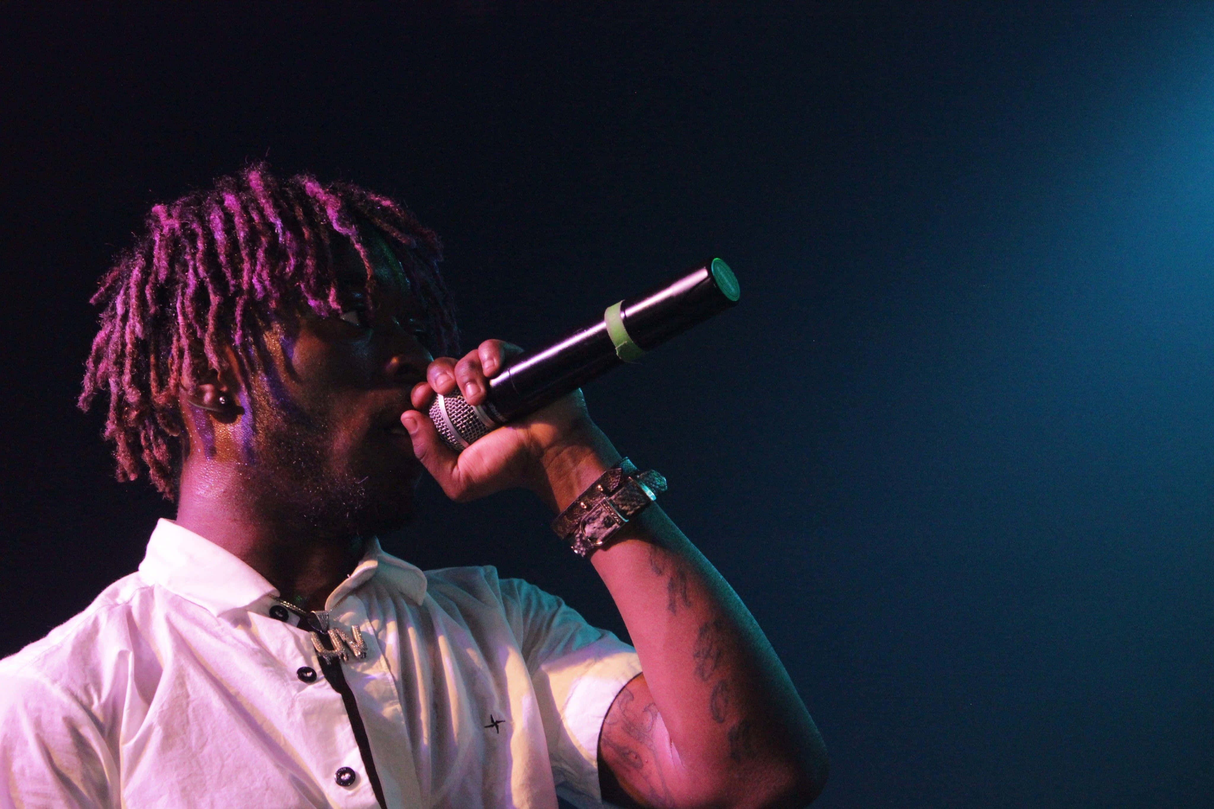 Lil Uzi Vert 桌布 壁紙 — 一位擁有紫色辮子頭的音樂家，在藍色舞台燈光下穿著白襯衫對著麥克風唱歌。