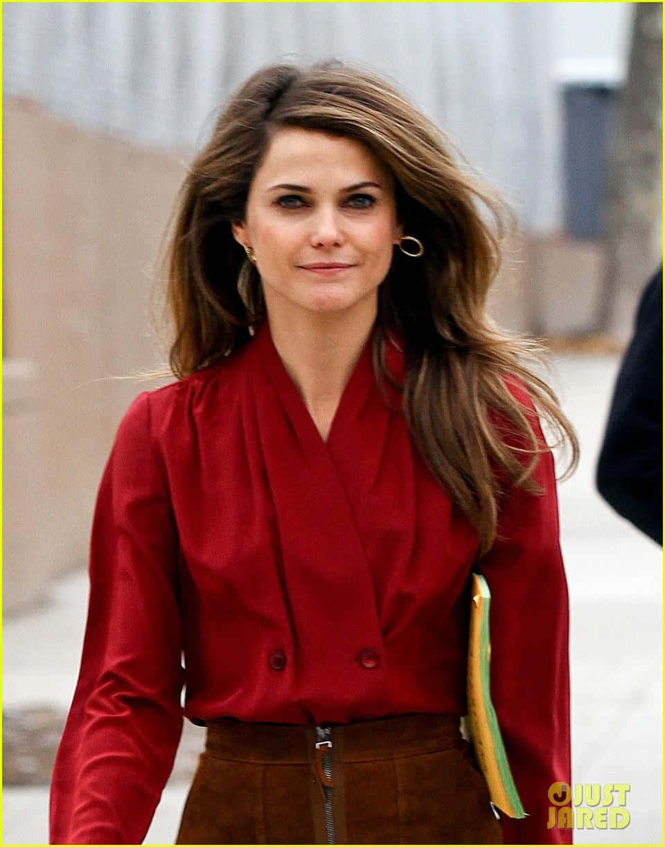 keri russell 桌布 壁紙 — 一位擁有被風吹亂棕色頭髮的女性，穿著大膽的紅色上衣和棕色褲子，在戶外行走。