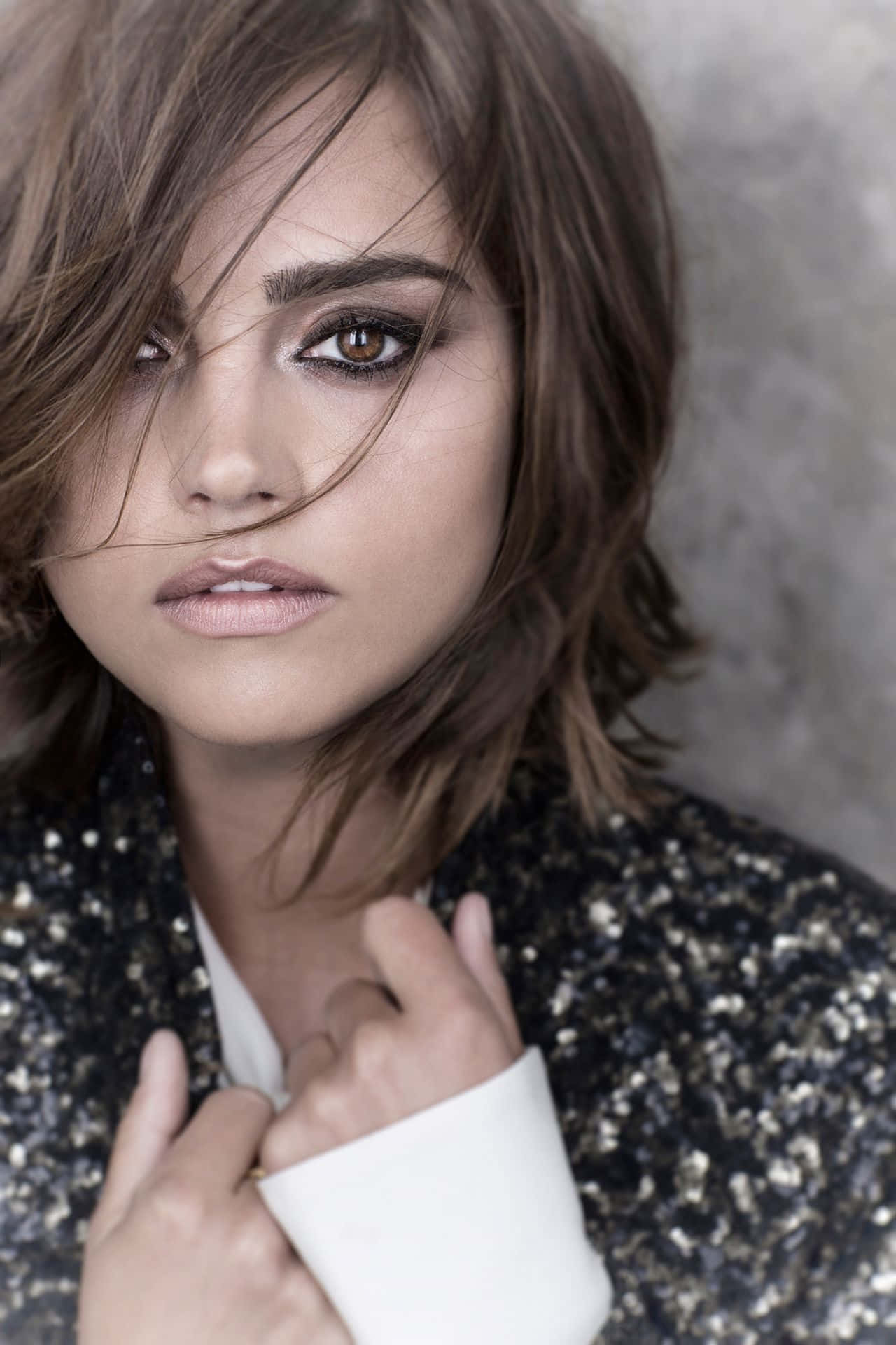 Jenna Coleman 桌布 壁紙 — 一位擁有被風吹亂的棕色頭髮和煙燻妝眼睛的女性，在紋理灰色牆壁前調整她的亮片外套，眼神強烈。