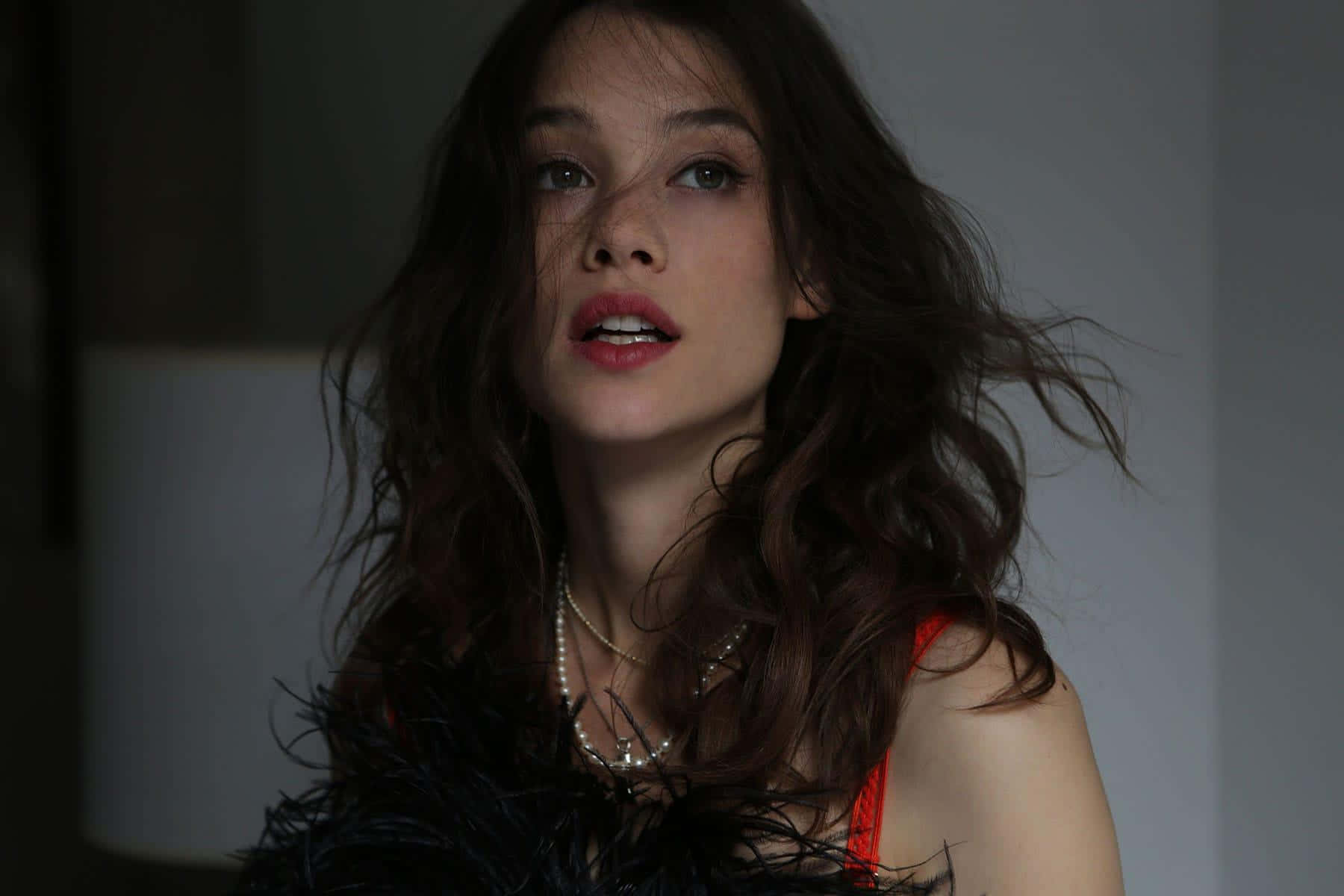 Astrid Berges Frisbey 桌布 壁紙 — 一位擁有被風吹亂的深色頭髮和紅唇的女性，在燈光昏暗的房間裡穿著羽毛上衣，向上凝視。