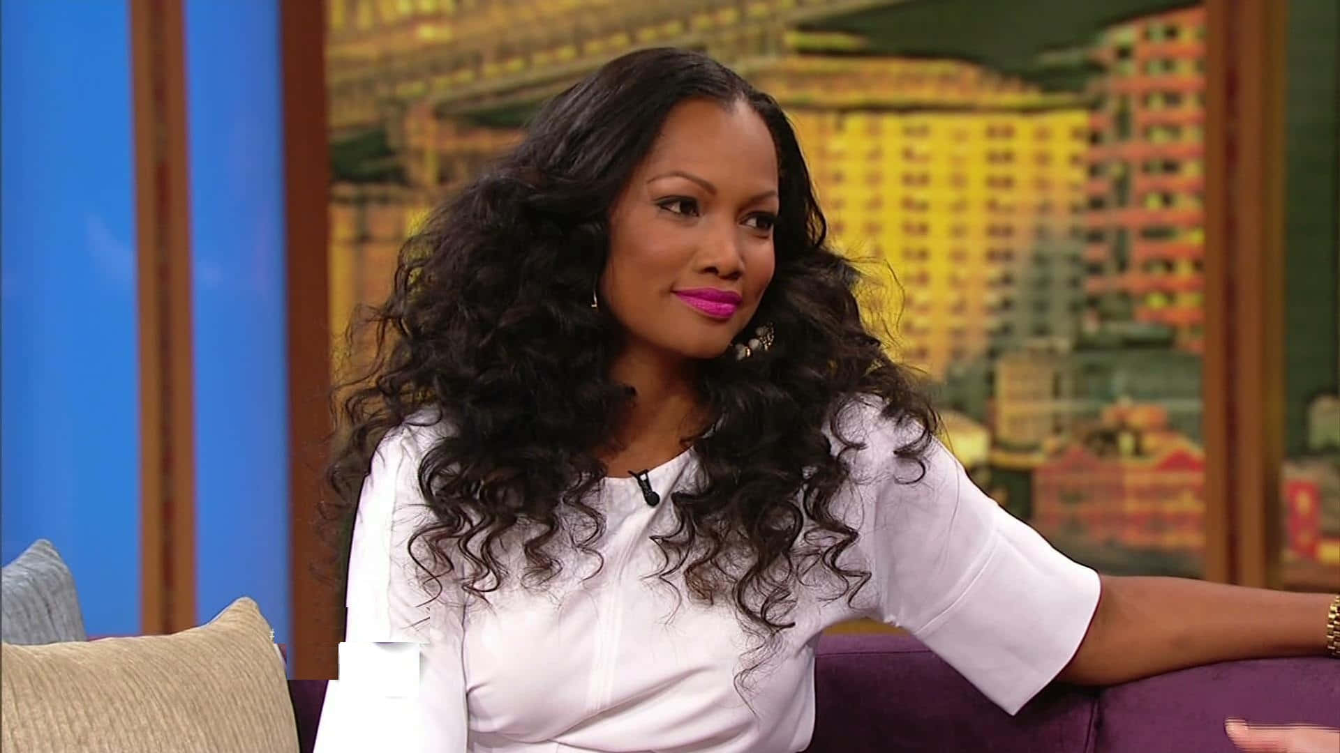 Garcelle Beauvais 桌布 壁紙 — 一位擁有豐盈深色捲髮和亮粉紅色唇膏的女性，坐在工作室的紫色沙發上，背景是城市天際線。