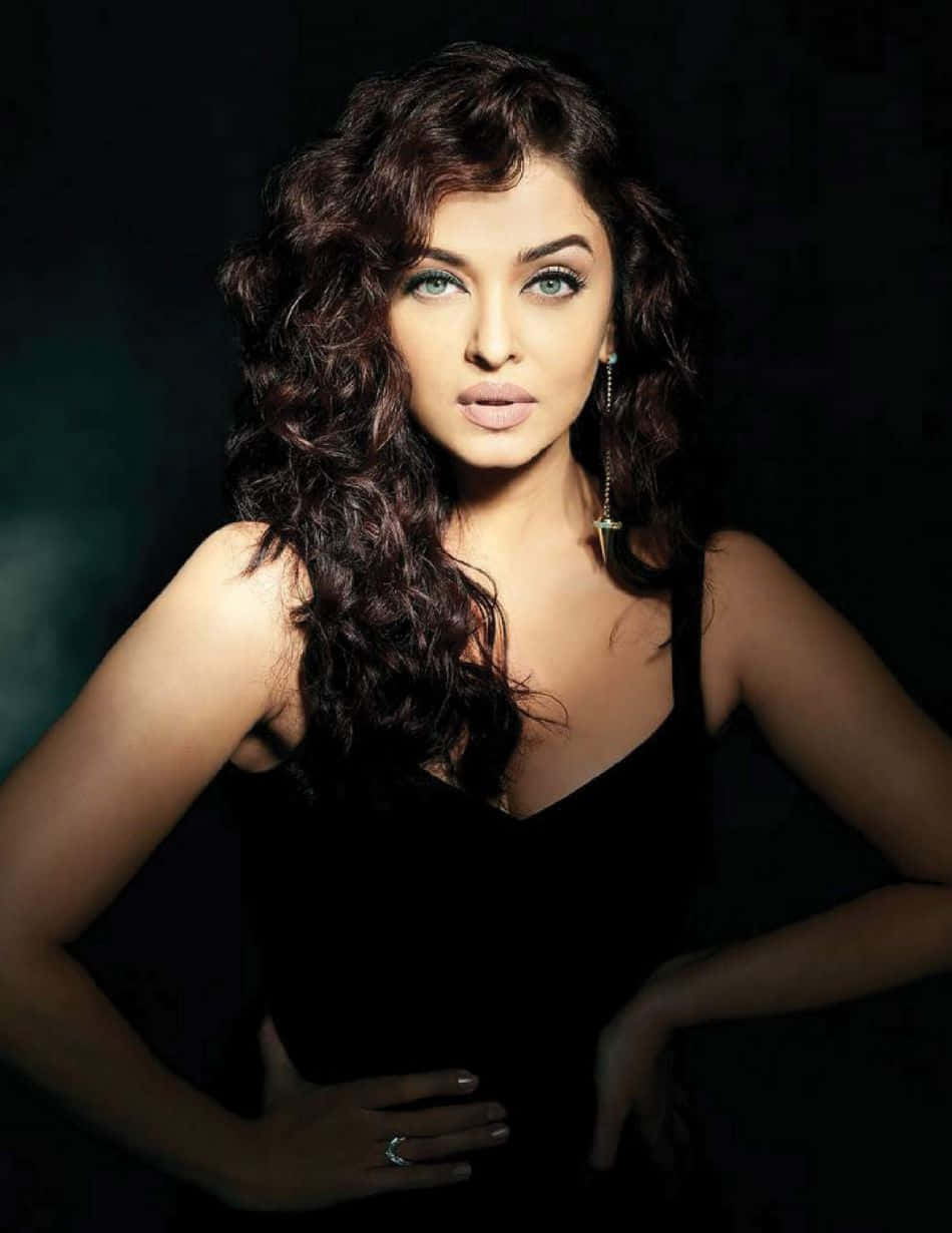 Aishwarya Rai 桌布 壁紙 — 一位擁有豐盈深色捲髮和引人注目的綠色眼睛的女性，在深色背景下自信地穿著黑色連衣裙擺姿勢。