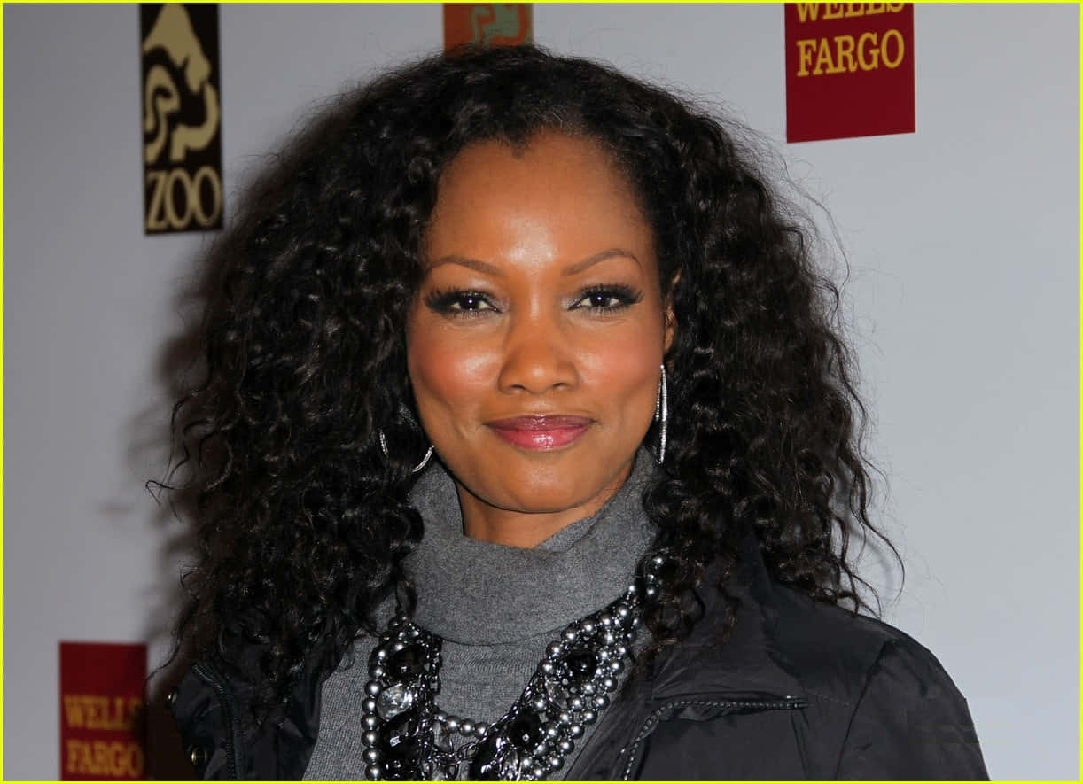 Garcelle Beauvais 桌布 壁紙 — 一位擁有豐盈深色捲髮的女性，穿著灰色高領毛衣和黑色外套，在紅地毯活動上對著鏡頭溫暖微笑。