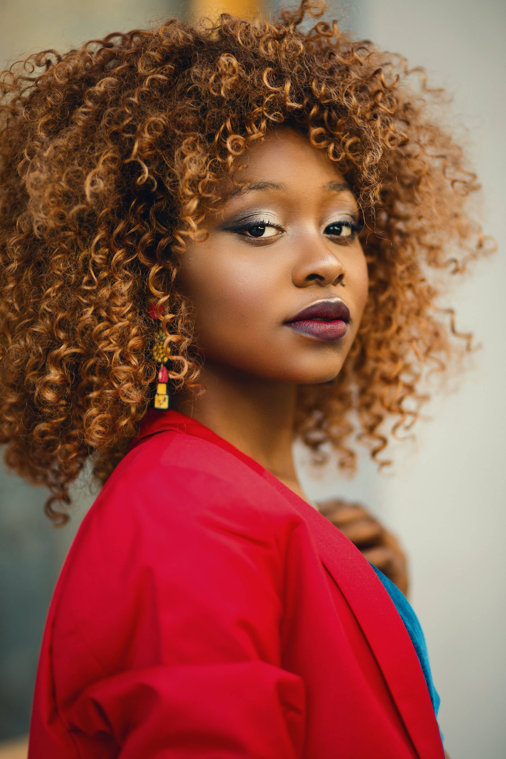black woman brown afro style 壁紙 — 一位擁有豐盈焦糖色捲髮、身穿大膽紅色外套的女性自信地凝視鏡頭。