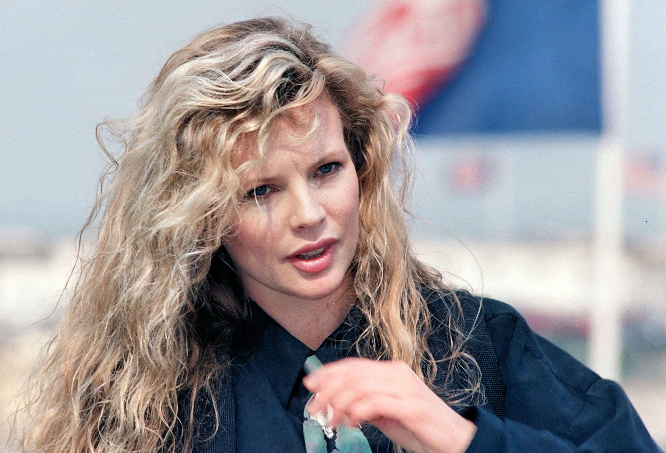 Kim Basinger At A 1989 Film Fest 壁紙 — 一位擁有豐盈金色捲髮、身穿深色西裝外套的女性，在模糊的城市背景下若有所思地凝視著。