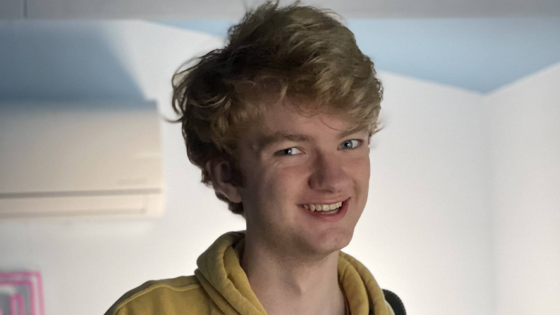 Thomas Simons Tommyinnit 壁紙 — 一位擁有金色捲髮和明亮藍眼睛的年輕男子，在室內穿著黃色連帽衫，露出溫暖的笑容。
