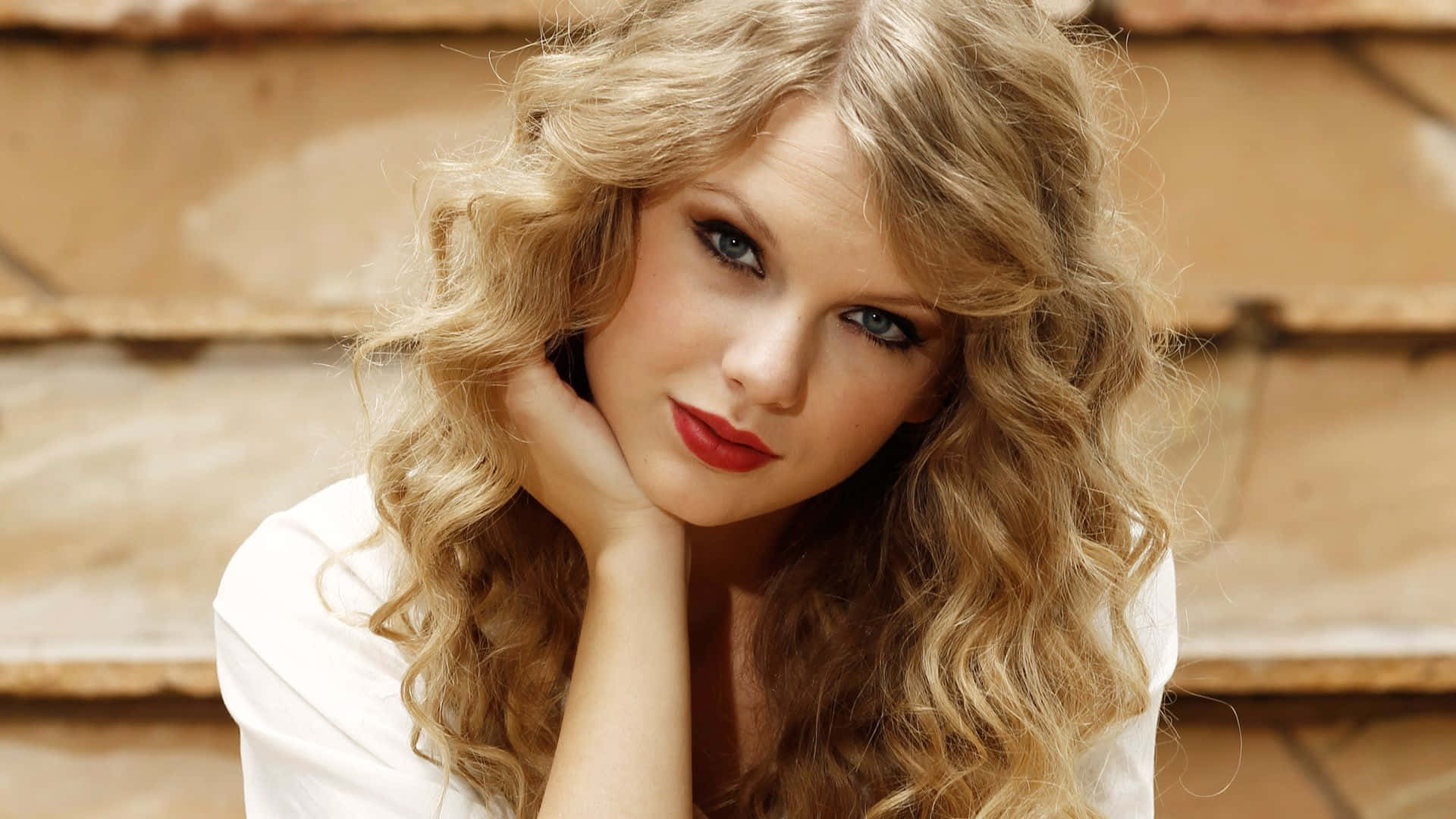 Taylor Swift 桌布 壁紙 — 一位擁有金色捲髮和紅唇的女性，將下巴擱在手上，靠著溫暖的石牆。