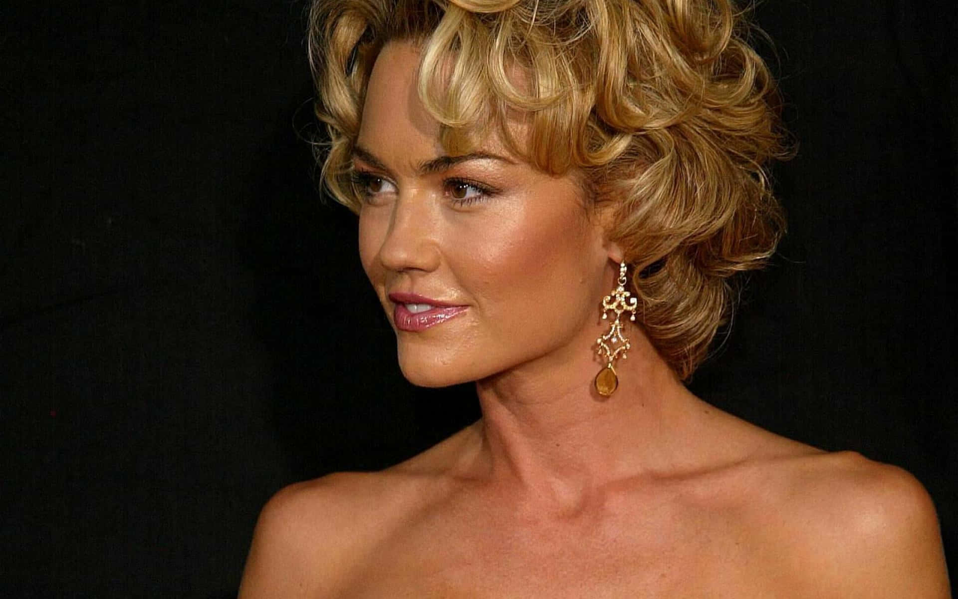 Kelly Carlson 桌布 壁紙 — 一位擁有金色捲髮和華麗耳環的魅力女性，在純黑背景前擺出姿勢。