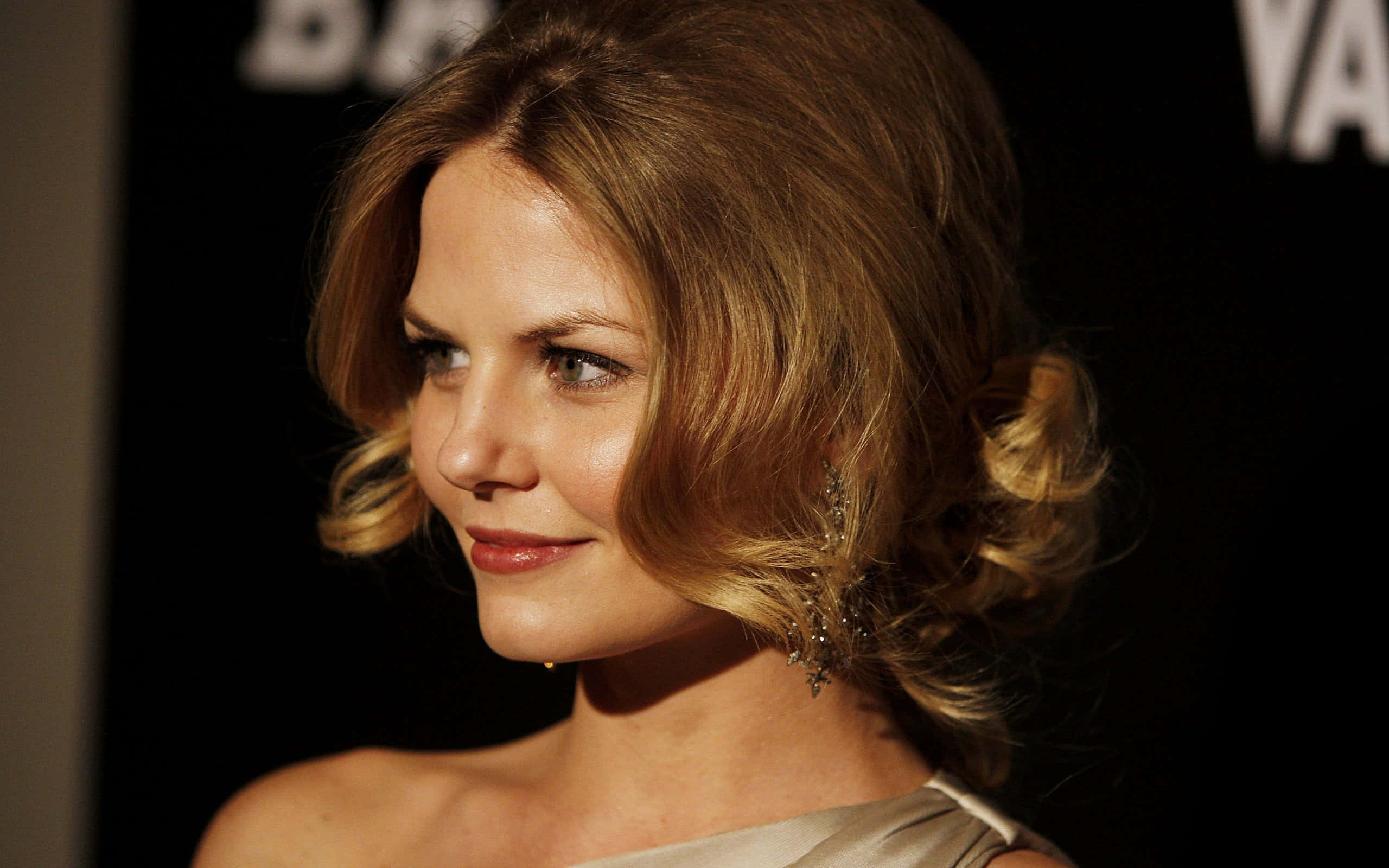 jennifer morrison 桌布 壁紙 — 一位擁有金色波浪髮型和優雅耳環的女性，在帶有模糊文字的深色背景下溫柔微笑。