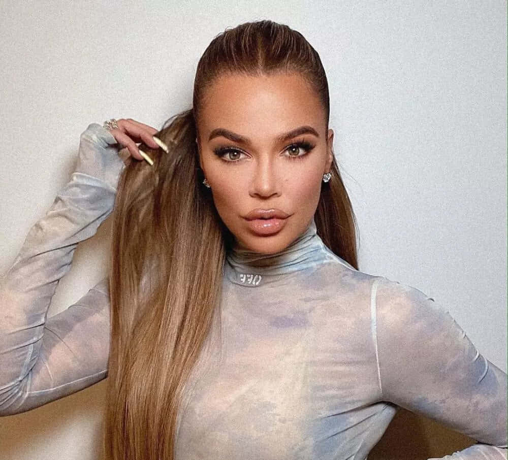 Khloe Kardashian 桌布 壁紙 — 一位擁有金色長髮和濃妝的女性，穿著透視銀色上衣，靠著素牆擺姿勢。