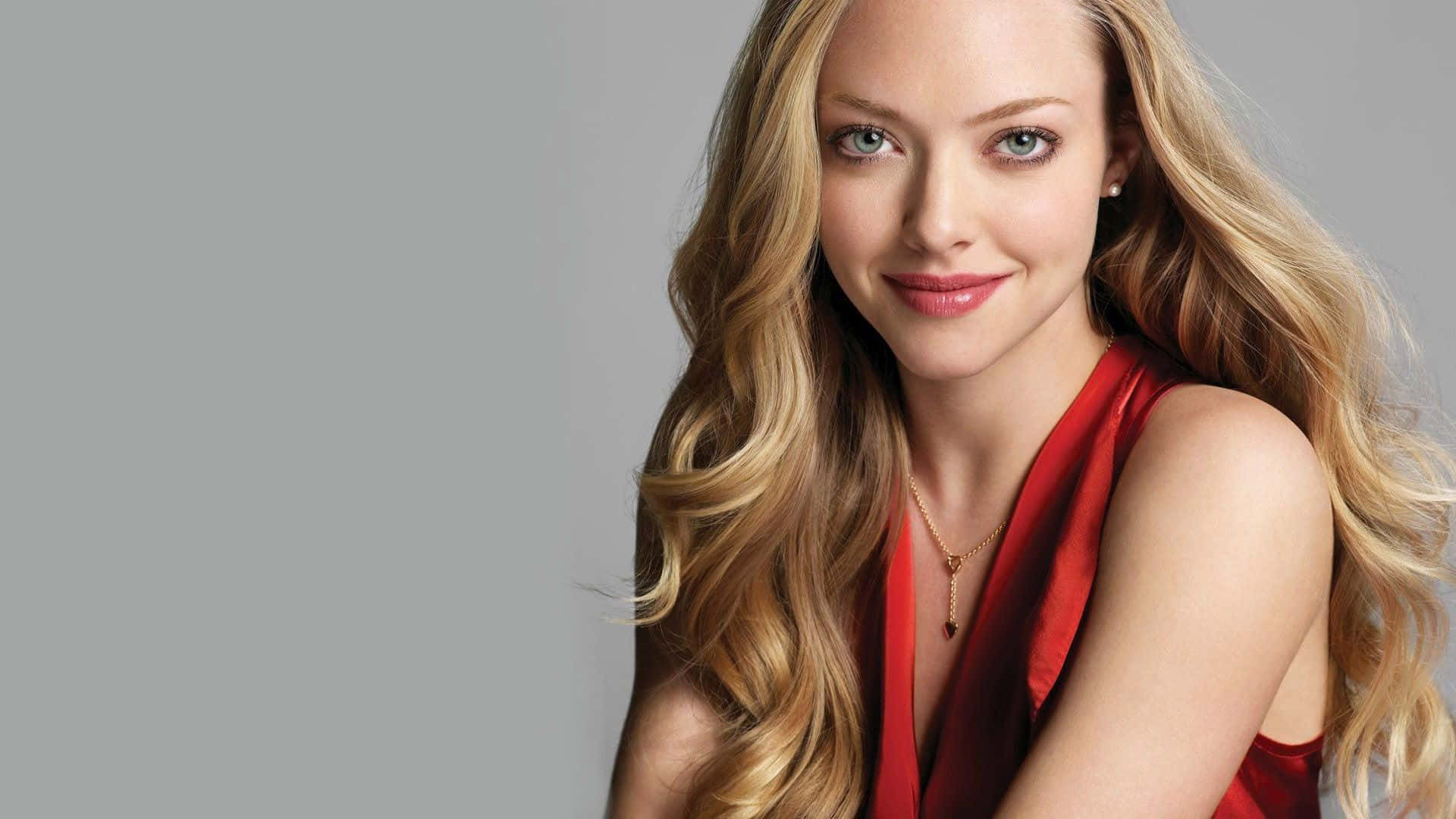 Amanda Seyfried 桌布 壁紙 — 一位擁有金色長髮和藍色眼睛的女性，穿著紅色緞面上衣，在純灰色背景前溫柔微笑。