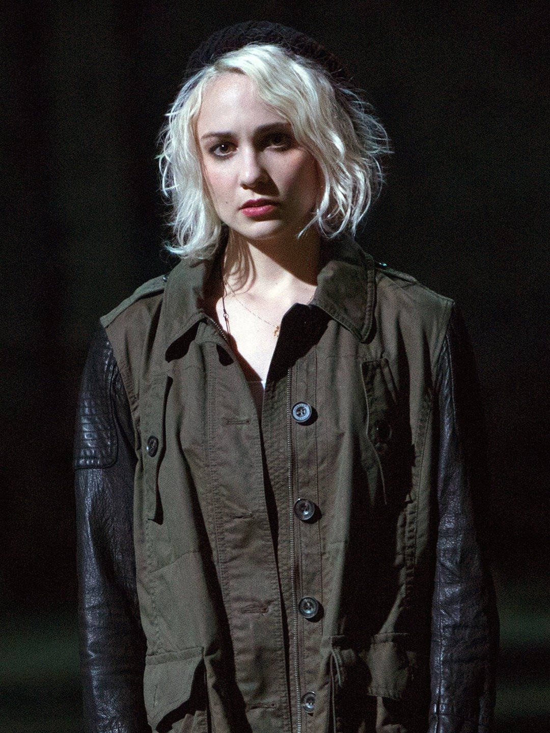 《Sense8》Riley Blue 壁紙 — 一位擁有鉑金色頭髮和黑色貝雷帽的女性，在昏暗的場景中穿著棕色背心搭配皮革袖子，專注地凝視著。
