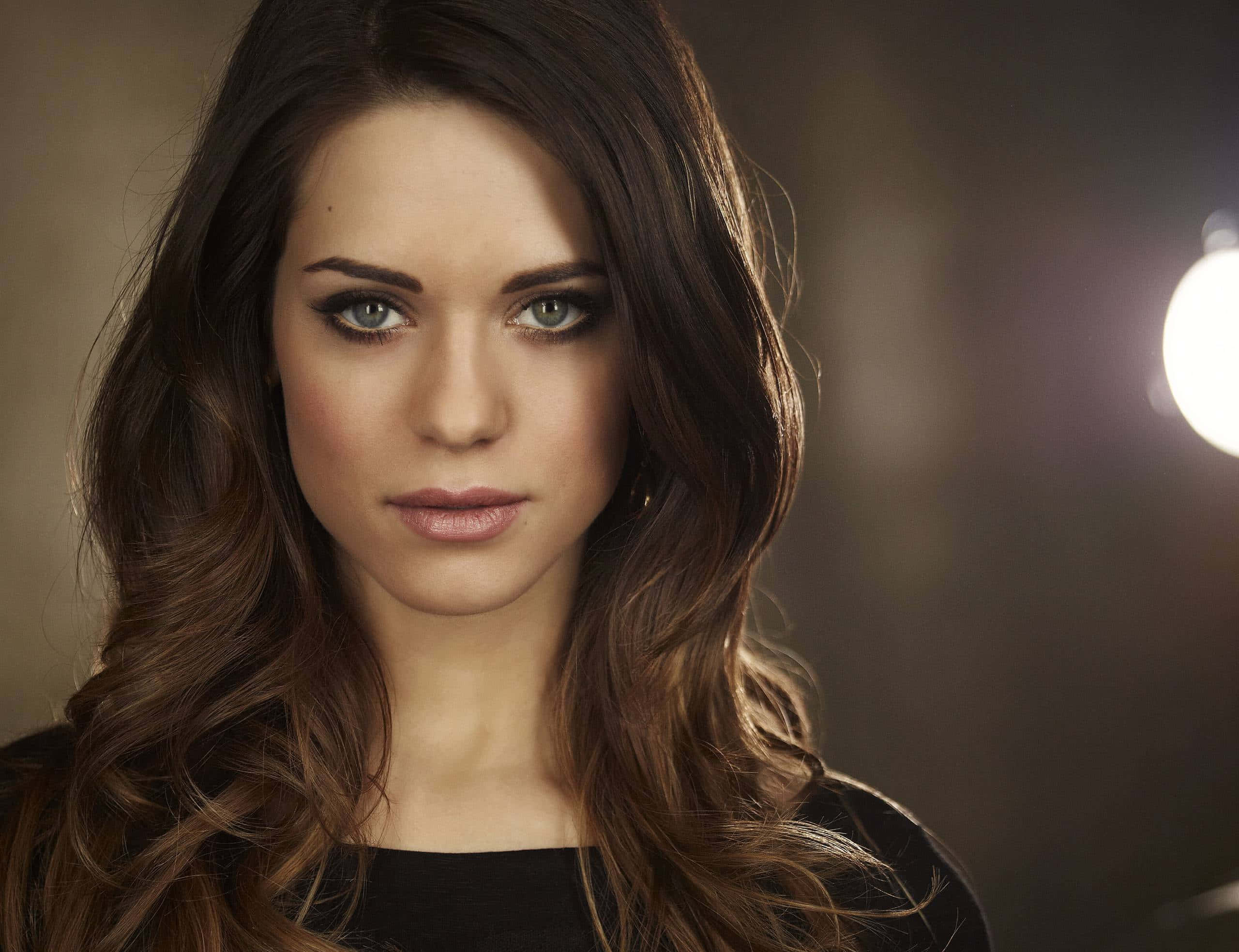lyndsy fonseca 桌布 壁紙 — 一位擁有長波浪棕色頭髮和引人注目的綠色眼睛的女性，在柔和的工作室燈光下直視鏡頭。