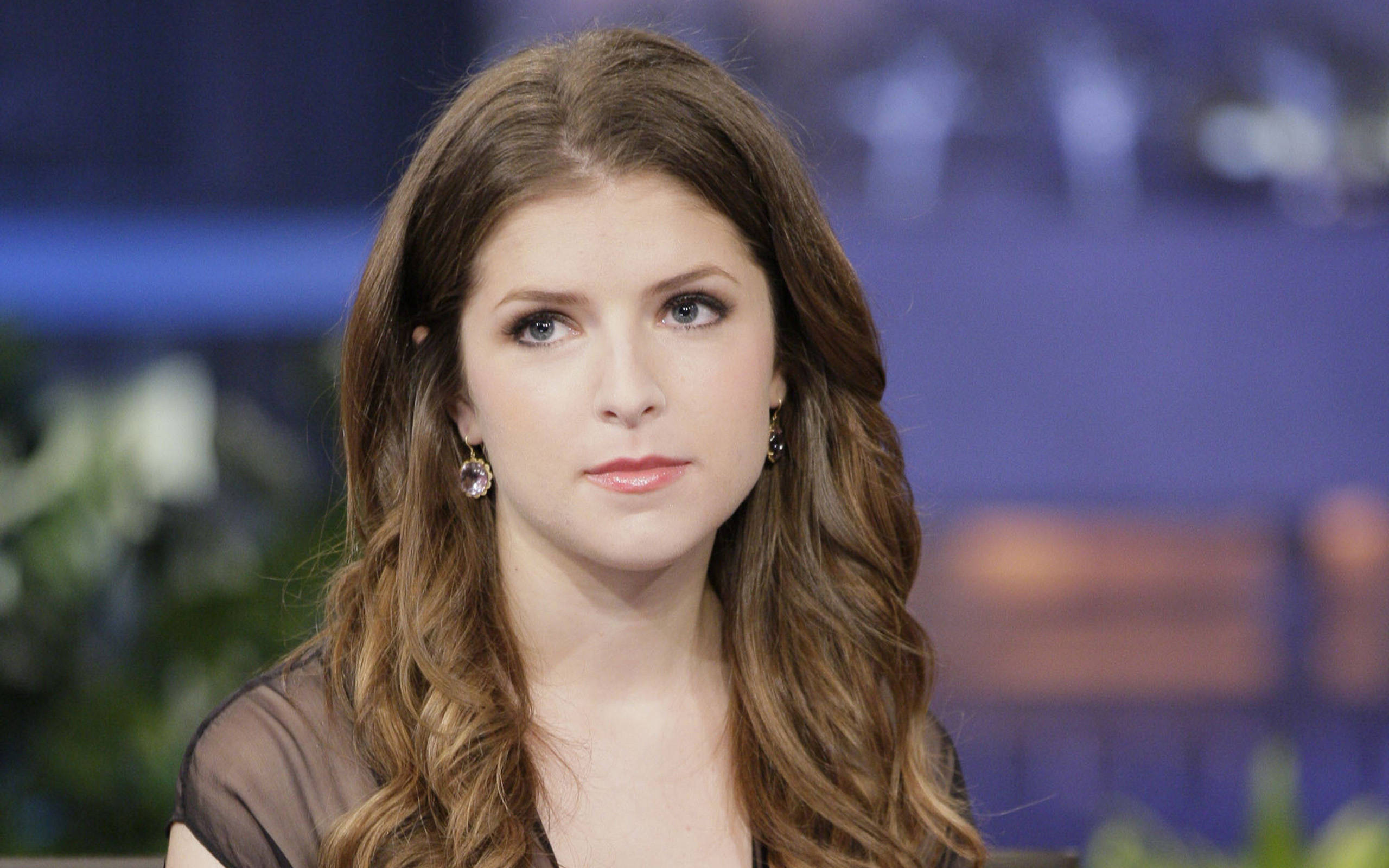 Cute Brunette Actress Anna Kendrick 壁紙 — 一位擁有長波浪棕色頭髮和藍色眼睛的女性，戴著垂墜耳環，若有所思地凝視著。