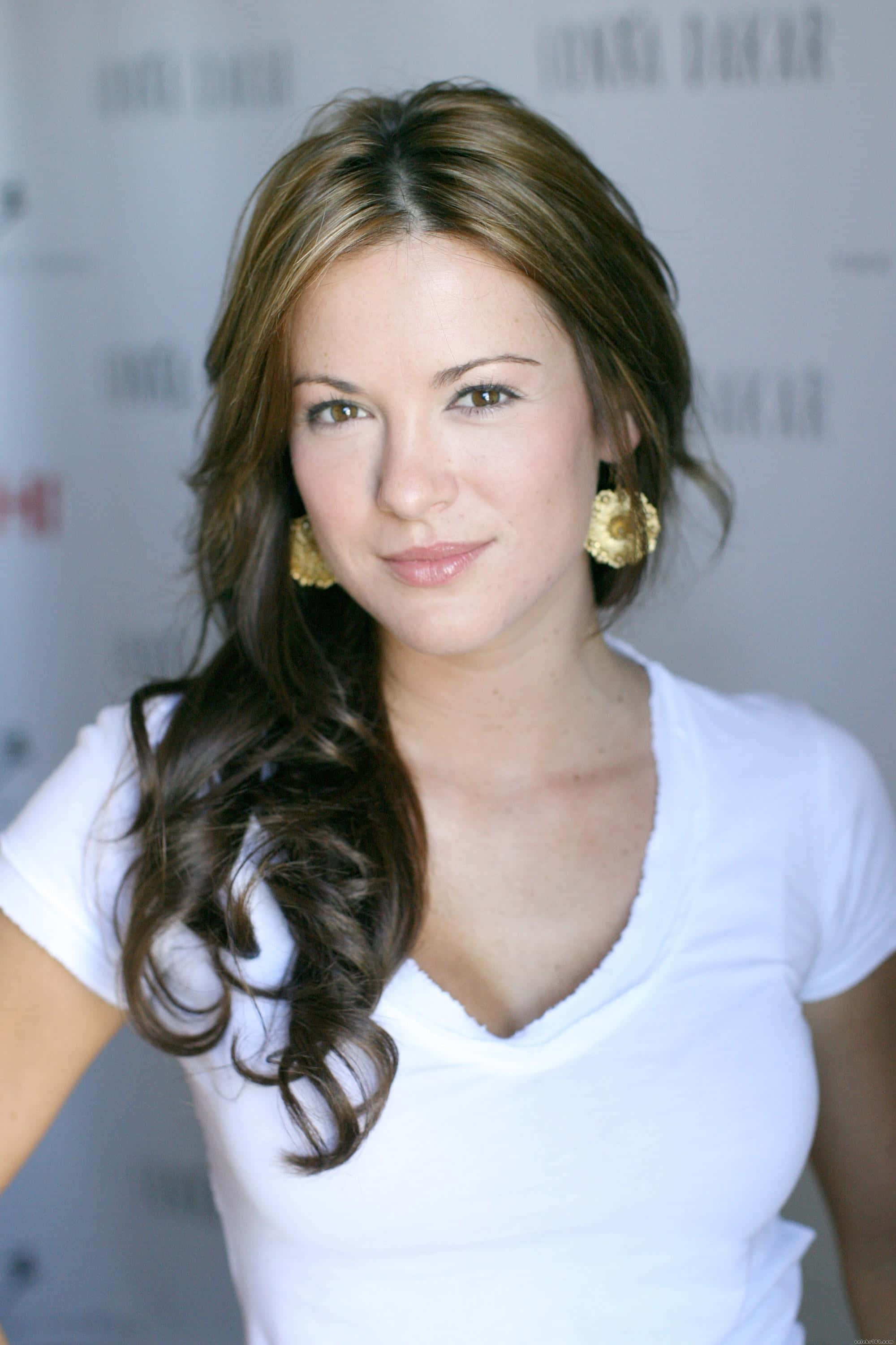 Danneel Ackles 桌布 壁紙 — 一位擁有長波浪棕色頭髮和金色耳環的女性，在品牌背景前穿著白色V領T恤，溫柔地微笑。